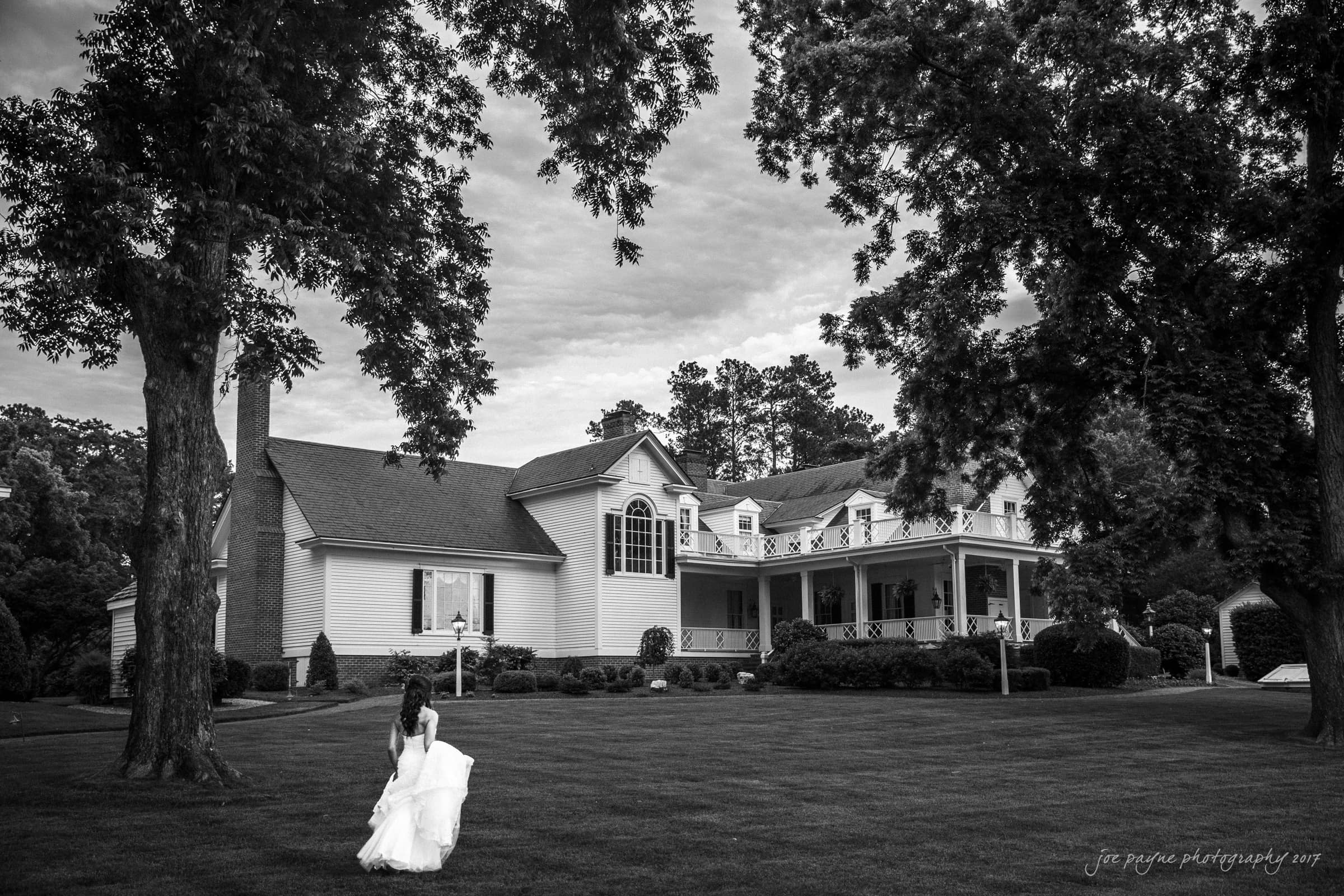 A bride in a flowing white gown stands on a manicured lawn, gazing at a large, elegant house with a wraparound porch and tall trees. The scene is shot in black and white, creating a timeless and serene atmosphere.