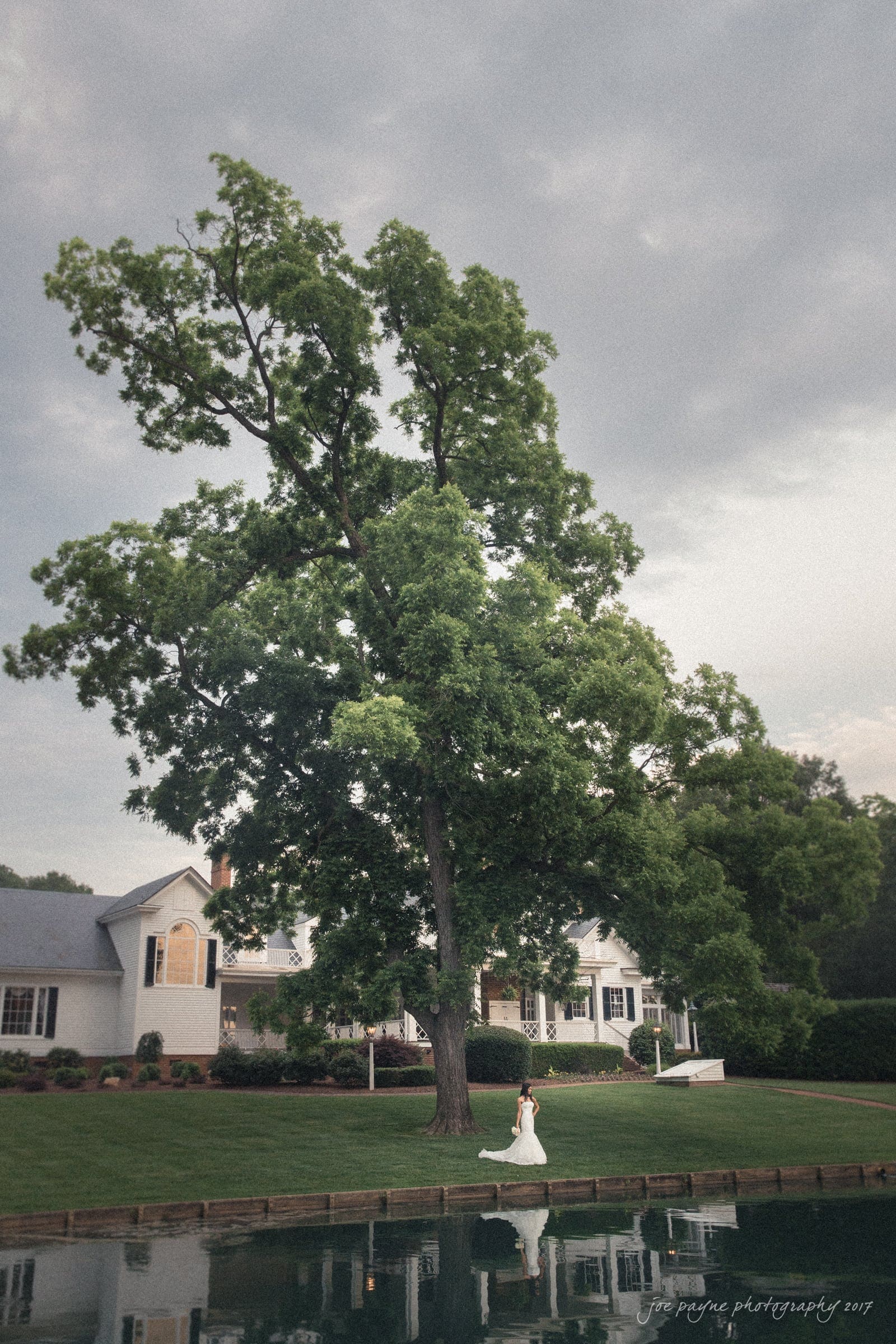 A bride in a white gown is walking along a pathway beside a large tree and a tranquil pond. A white house with a porch and manicured lawn is in the background under a cloudy sky.