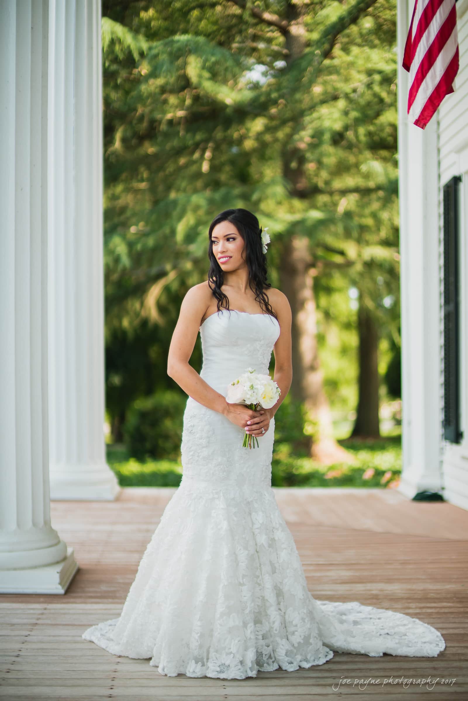 A bride in a white, strapless lace gown holding a bouquet of white flowers stands on a wooden porch. She is surrounded by white columns and greenery, with an American flag visible in the corner.