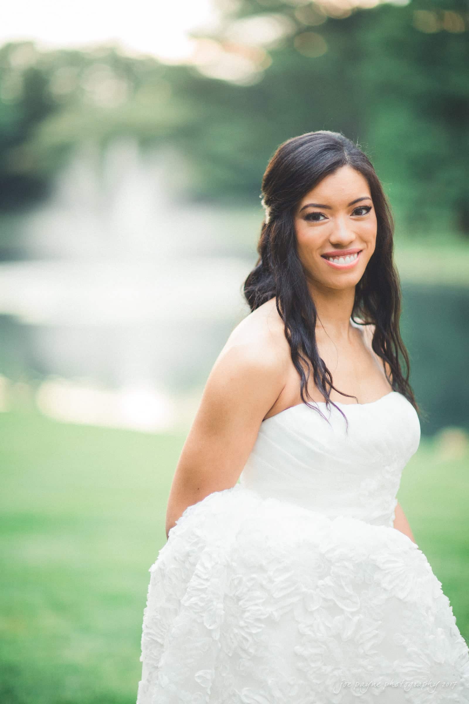 A bride in a white dress smiles outdoors with a blurred background of greenery and a pond. Her dark hair is styled in loose waves. The sunlight softly illuminates the scene, creating a serene and joyful atmosphere.