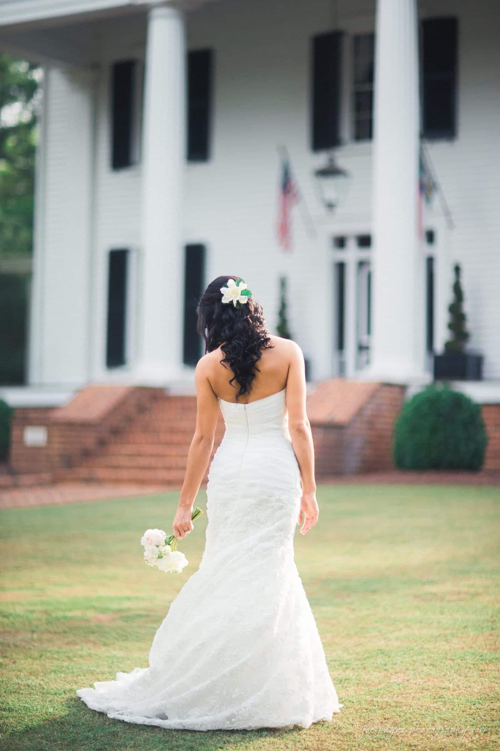 A bride in a strapless white gown stands on a lawn, holding a bouquet, with her back to the camera. Her dark hair is styled with a flower accessory. In the background is a white building with columns and flags.