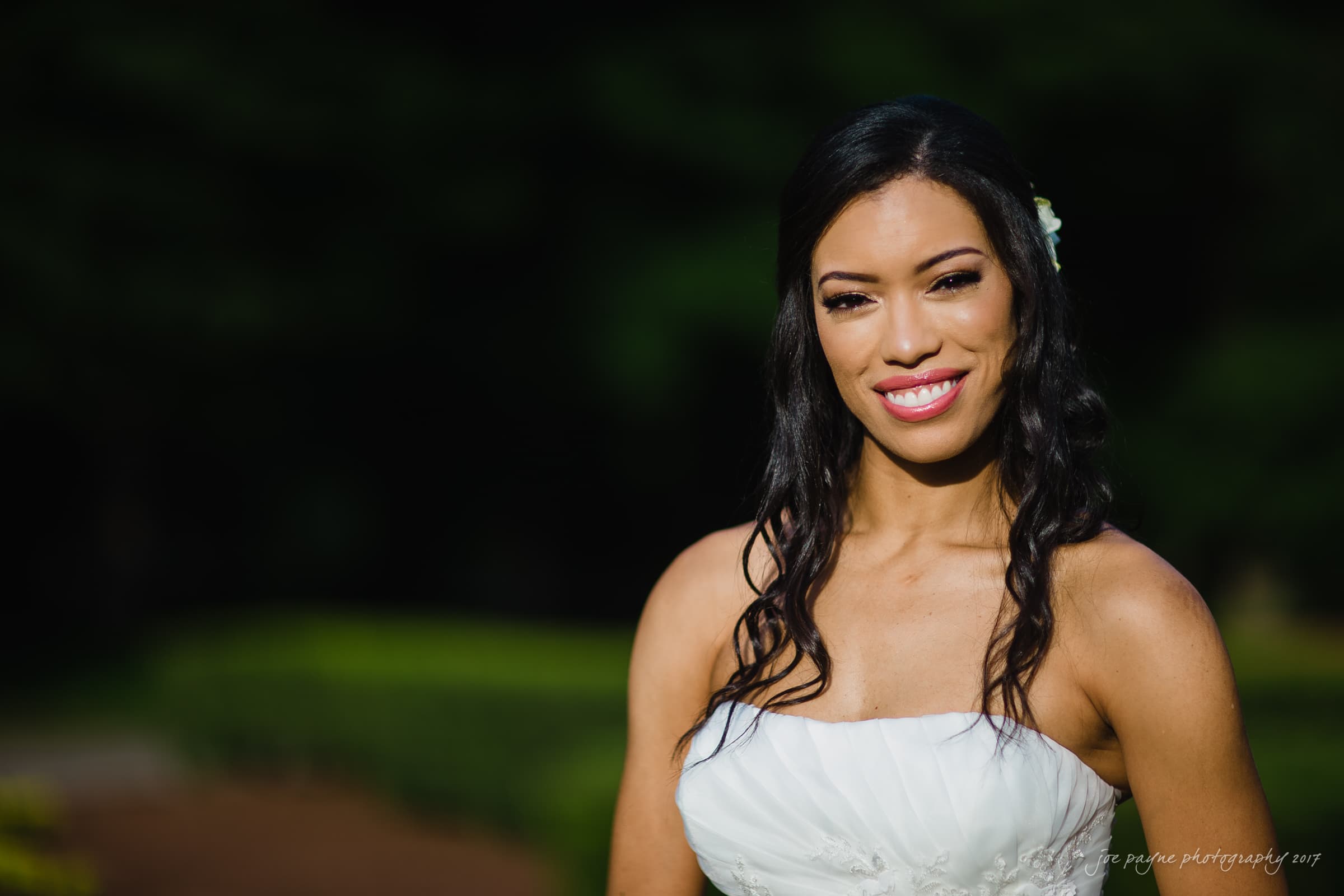 A woman in a white strapless dress smiles warmly at the camera. Her long dark hair falls in loose waves, and the background is a vibrant, blurred green, suggesting lush foliage on a sunny day.