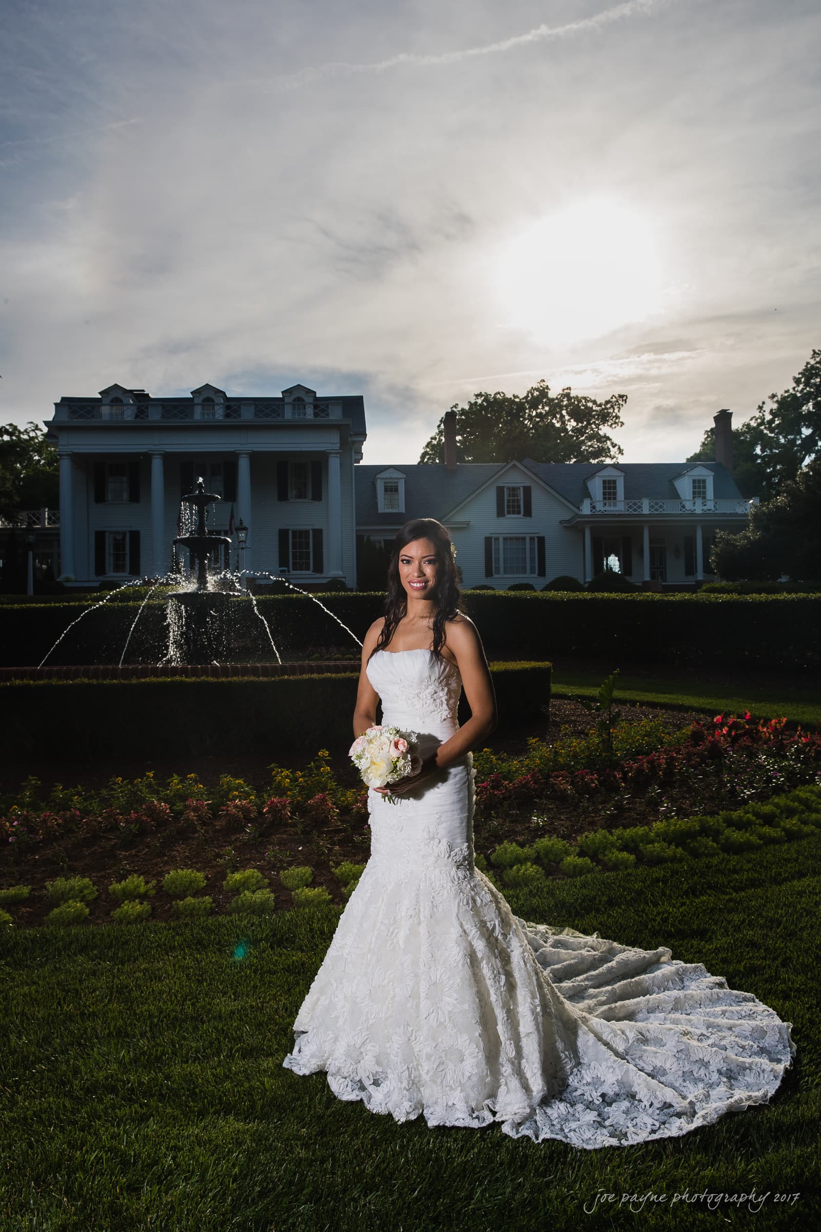Bride in a white lace gown stands in front of a large fountain and elegant house, holding a bouquet of white flowers. The sun sets in the background, illuminating the scene with a warm glow.
