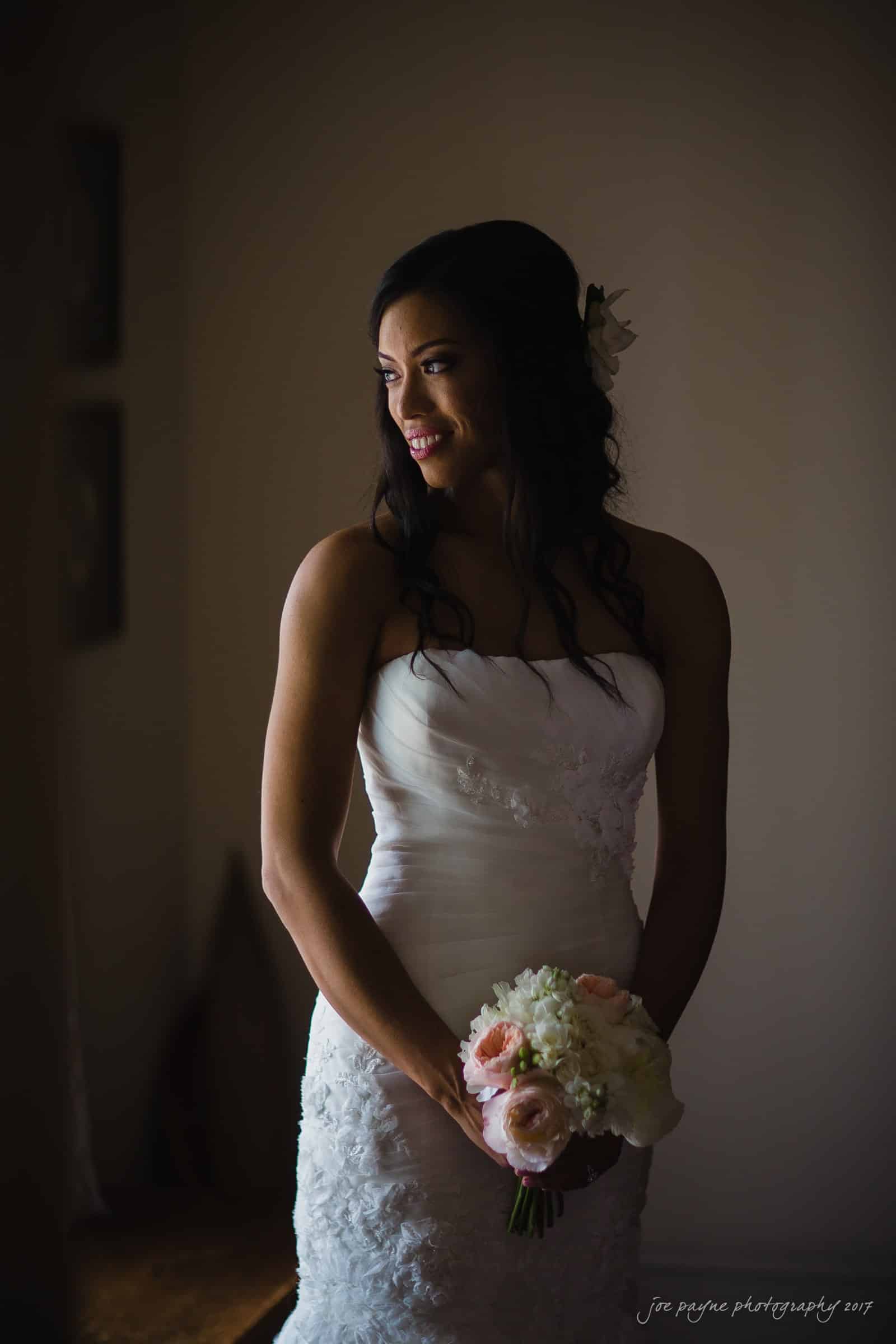 A bride in a white wedding dress stands near a window, holding a bouquet of white and pink flowers. Soft light illuminates her as she looks to the side, creating a serene and elegant atmosphere.