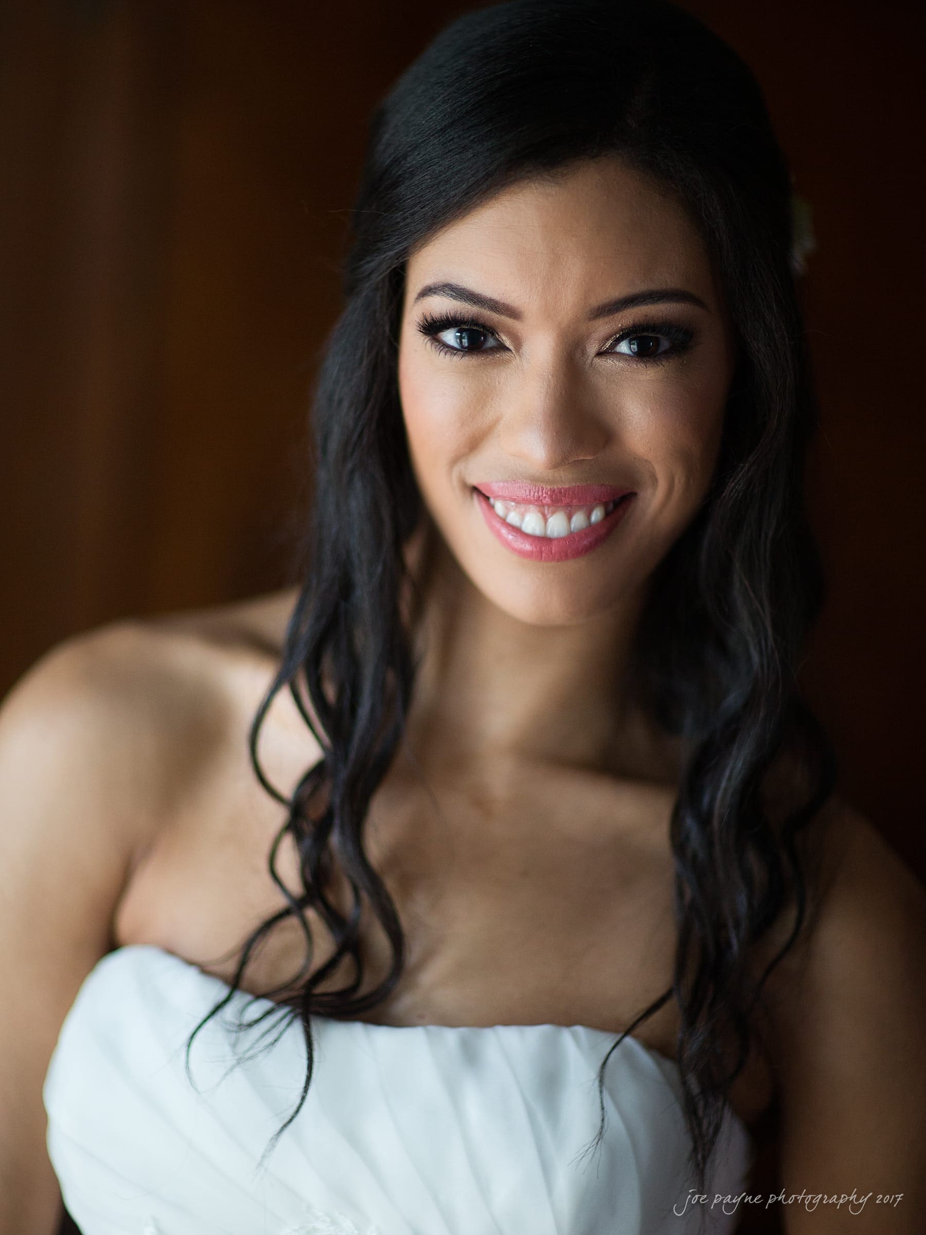 A woman with long, dark hair smiles directly at the camera. She is wearing a white strapless dress, and the background is softly blurred.