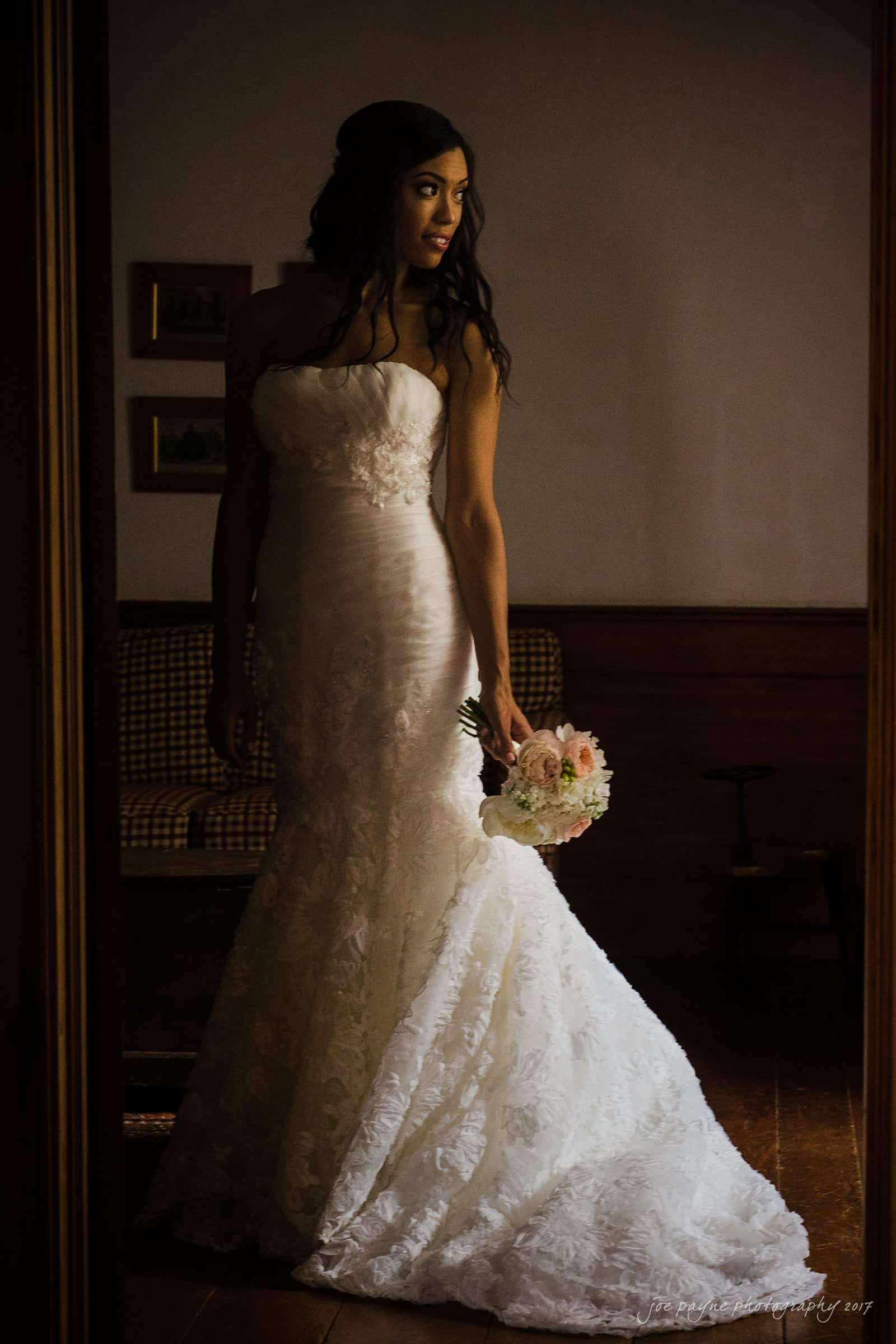 A bride in a strapless white lace gown holds a bouquet of flowers. She stands in a dimly lit room, looking to the side, with her long hair cascading over her shoulders. The setting is warm and intimate.