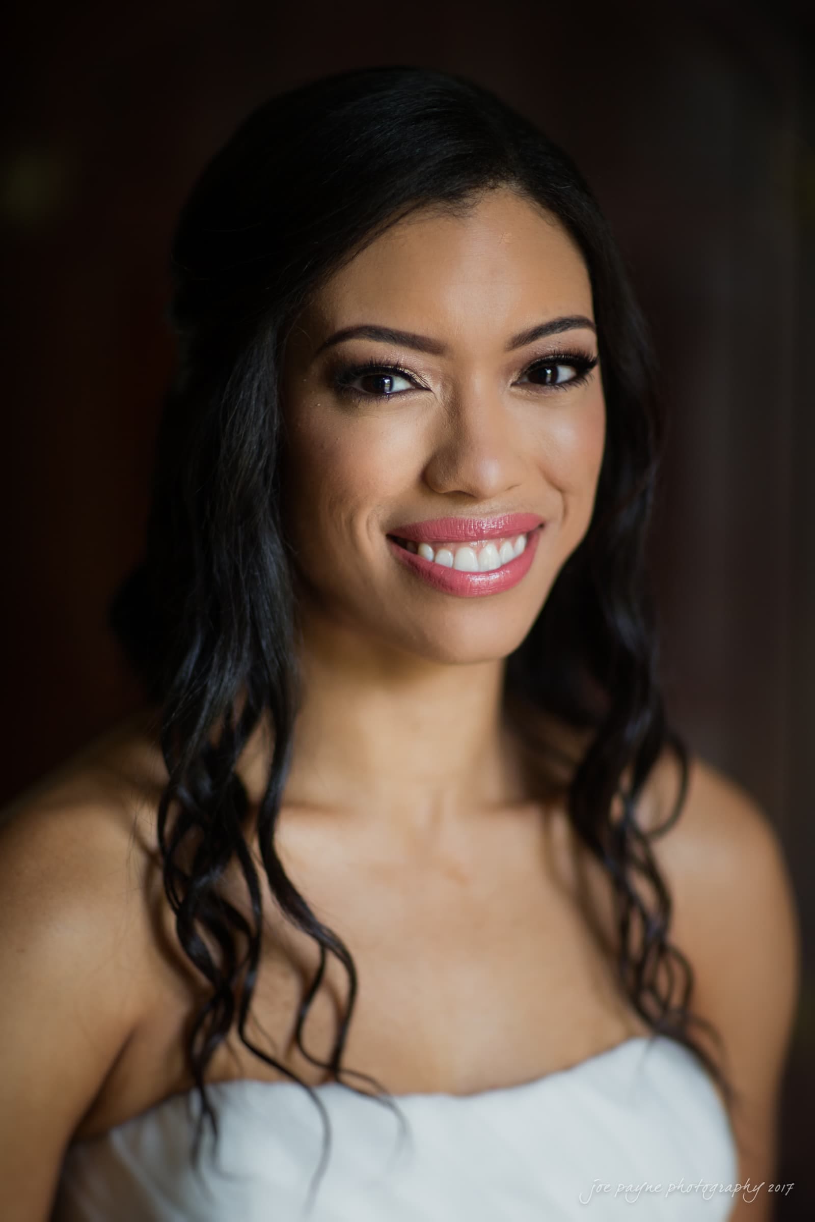 Smiling woman with long dark hair and subtle makeup wearing a white strapless top against a dark background.