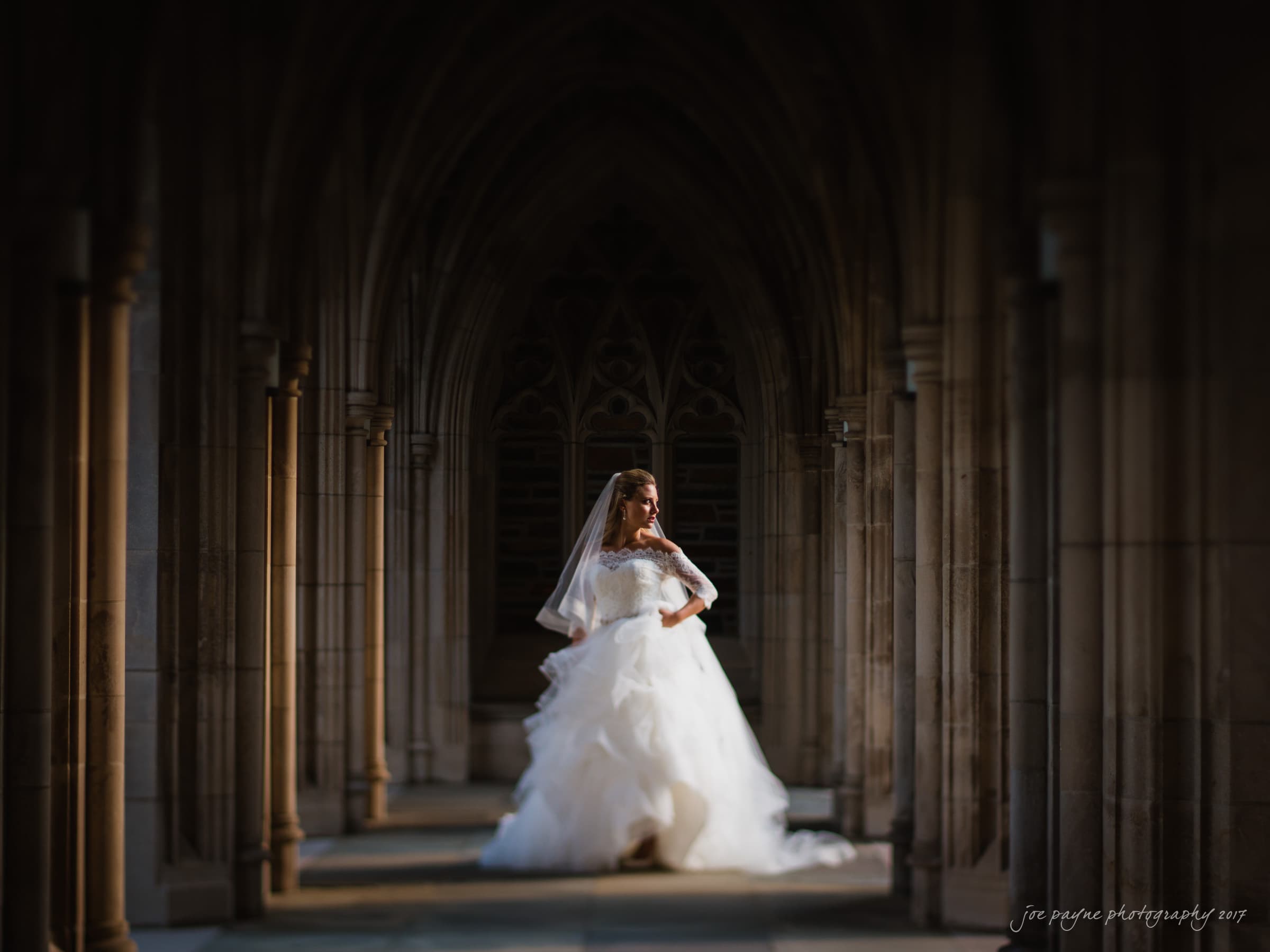 duke chapel wedding photography - sara's bridal session A bride in a white gown and veil stands in the center of a Gothic-style corridor with arched columns. Sunlight softly illuminates her and casts shadows in the architectural setting, creating a dramatic and elegant atmosphere.