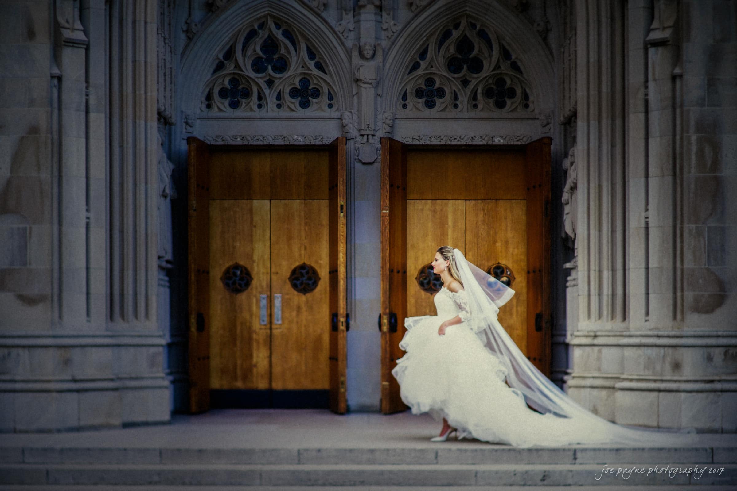duke chapel wedding photography - sara's bridal session A bride in a white gown and veil stands on stone steps in front of large wooden doors with intricate stone carvings above them. The setting appears to be a grand, historical building, adding a regal atmosphere to the scene.