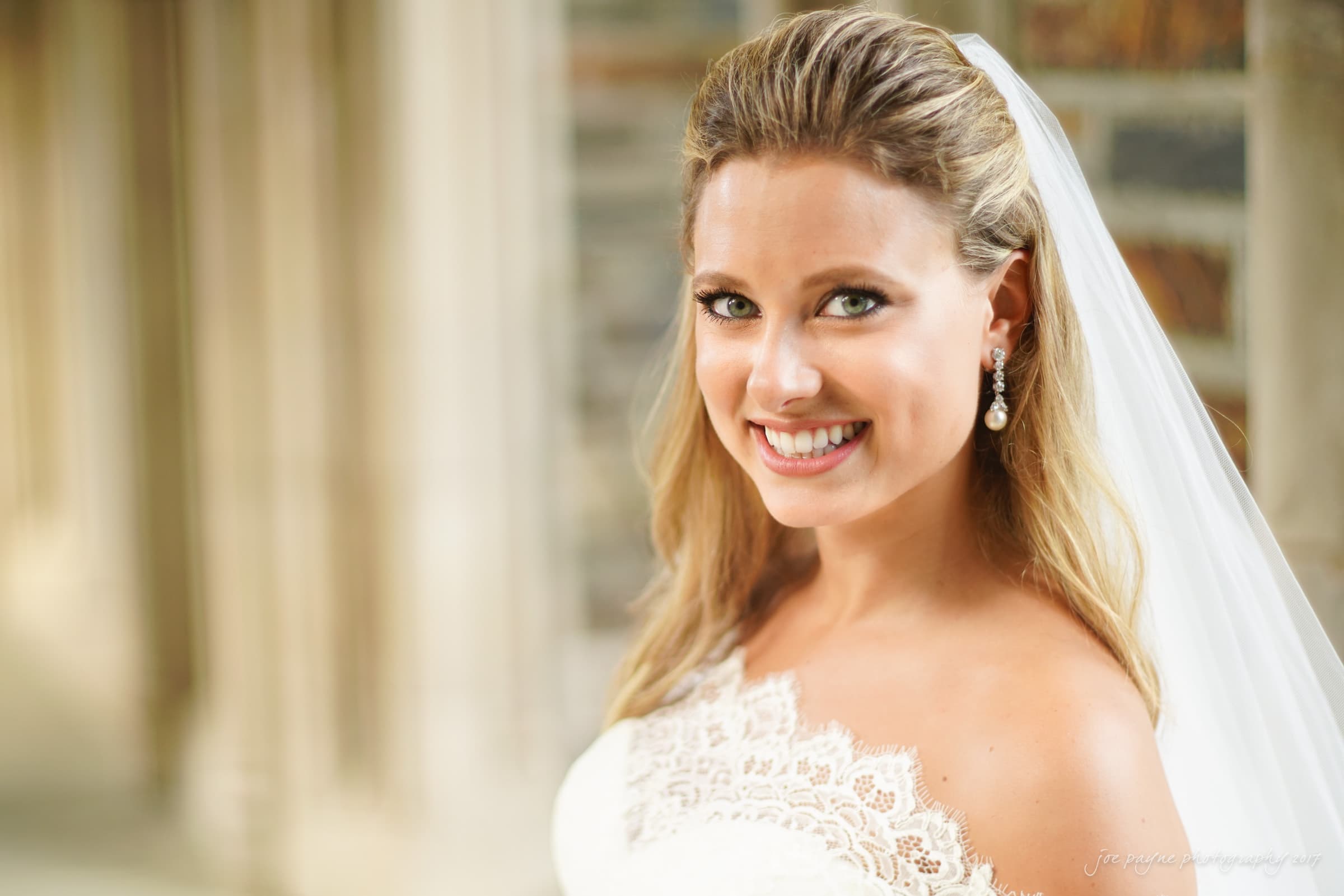 duke chapel wedding photography - sara's bridal session Smiling bride in a white lace dress and veil, standing near a stone building, wearing pearl earrings.