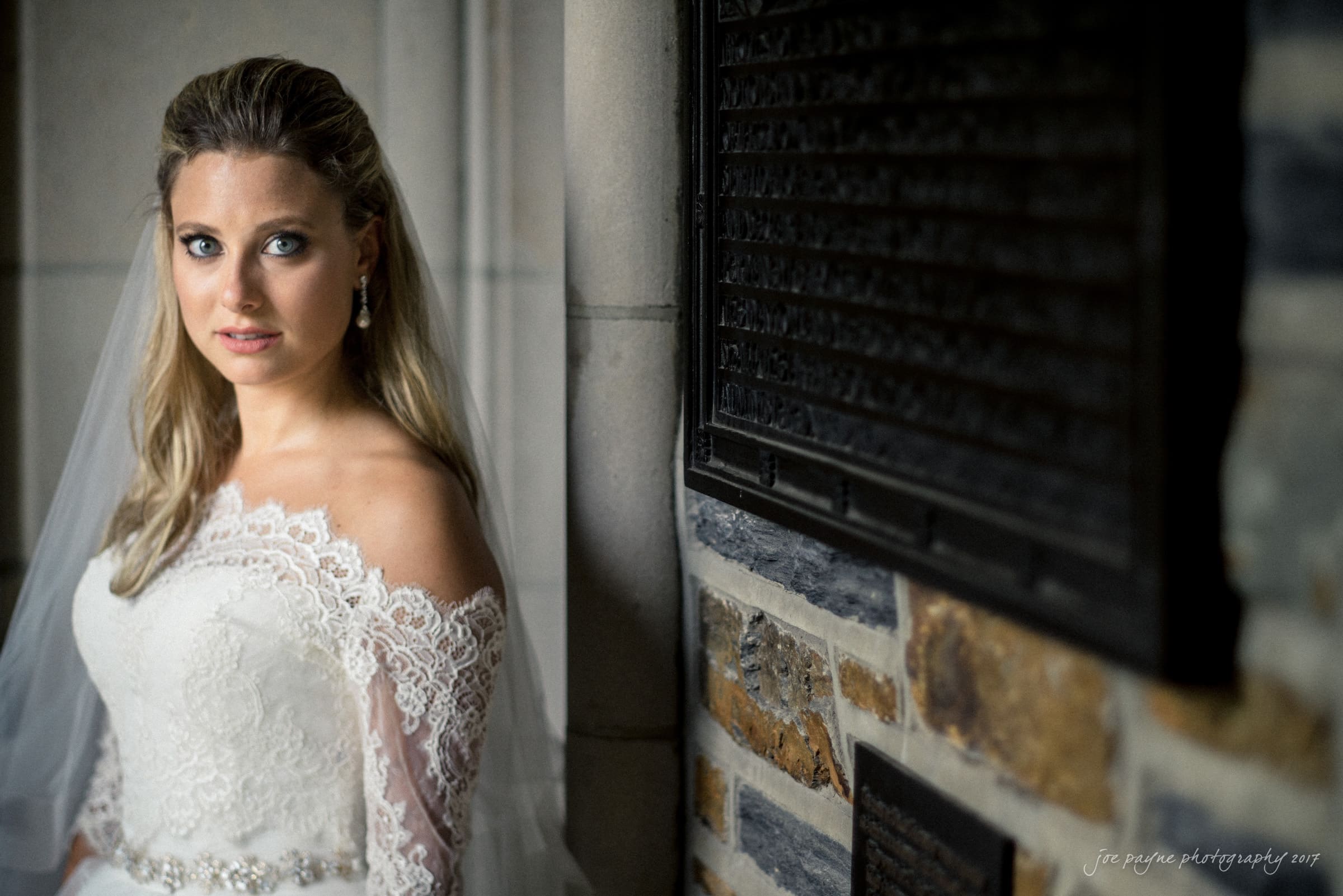 duke chapel wedding photography - sara's bridal session A bride in a white lace wedding dress with off-the-shoulder sleeves stands next to a stone wall. She has long, wavy hair and is wearing earrings. Her expression is calm as she gazes at the camera.