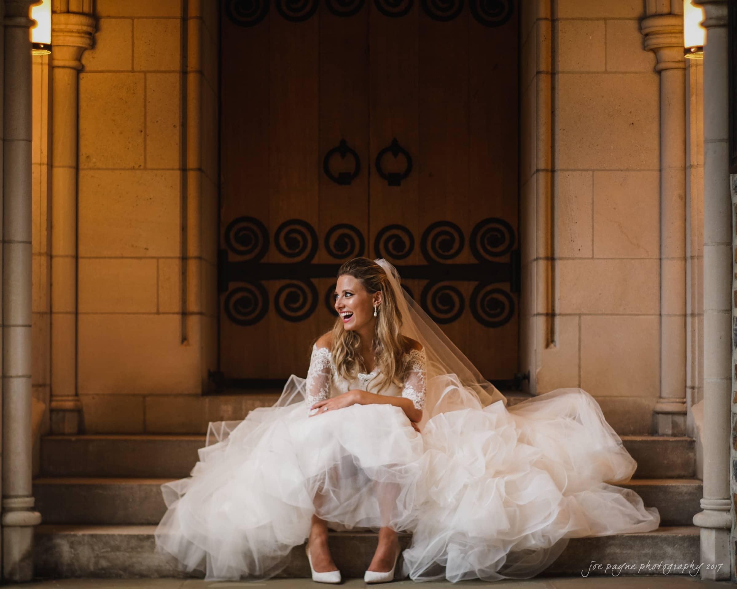 duke chapel wedding photography - sara's bridal session A bride in a white wedding dress sits on steps in front of wooden doors with intricate designs. She smiles joyfully, surrounded by the soft glow of warm lighting on either side of the entrance.