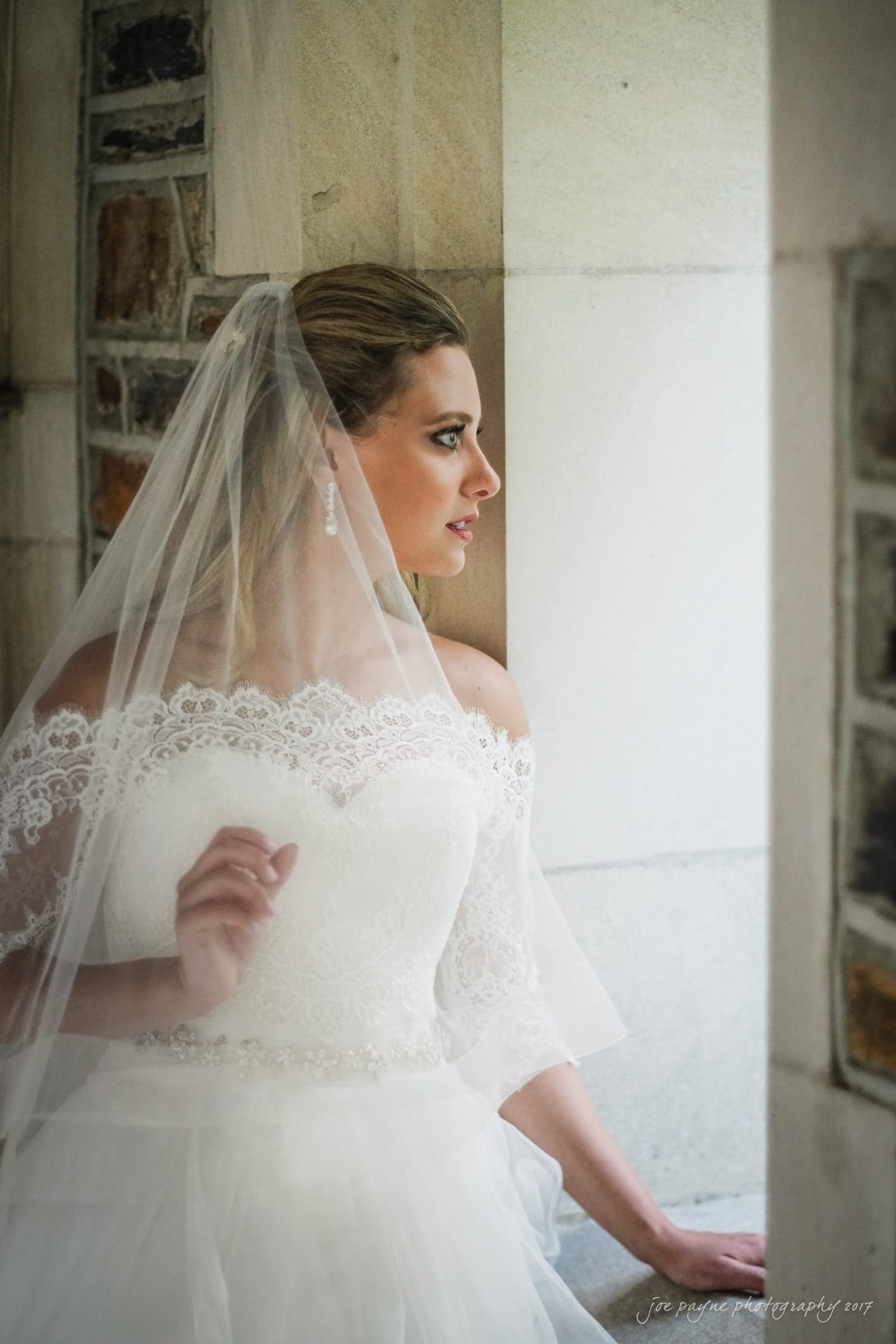 duke chapel wedding photography - sara's bridal session A bride in a lace off-the-shoulder wedding dress and veil stands by a window. She looks thoughtfully outward, her hand gently touching the windowsill. Stone walls and an archway are visible in the background.