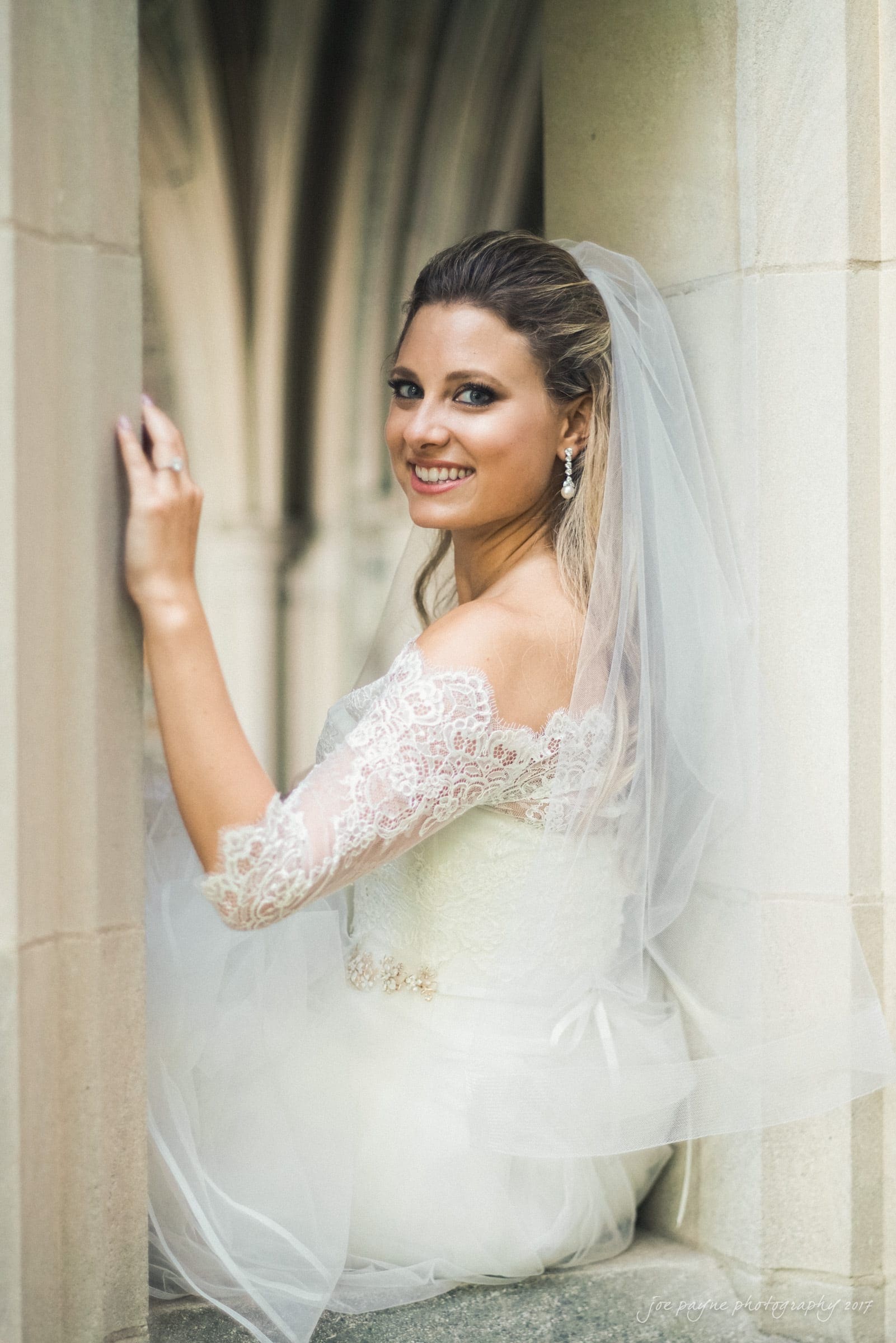 duke chapel wedding photography - sara's bridal session A bride in a white lace wedding dress and veil smiles while sitting on a stone ledge. She leans against a stone archway, with soft natural light illuminating her, showcasing her earrings and hairstyle.