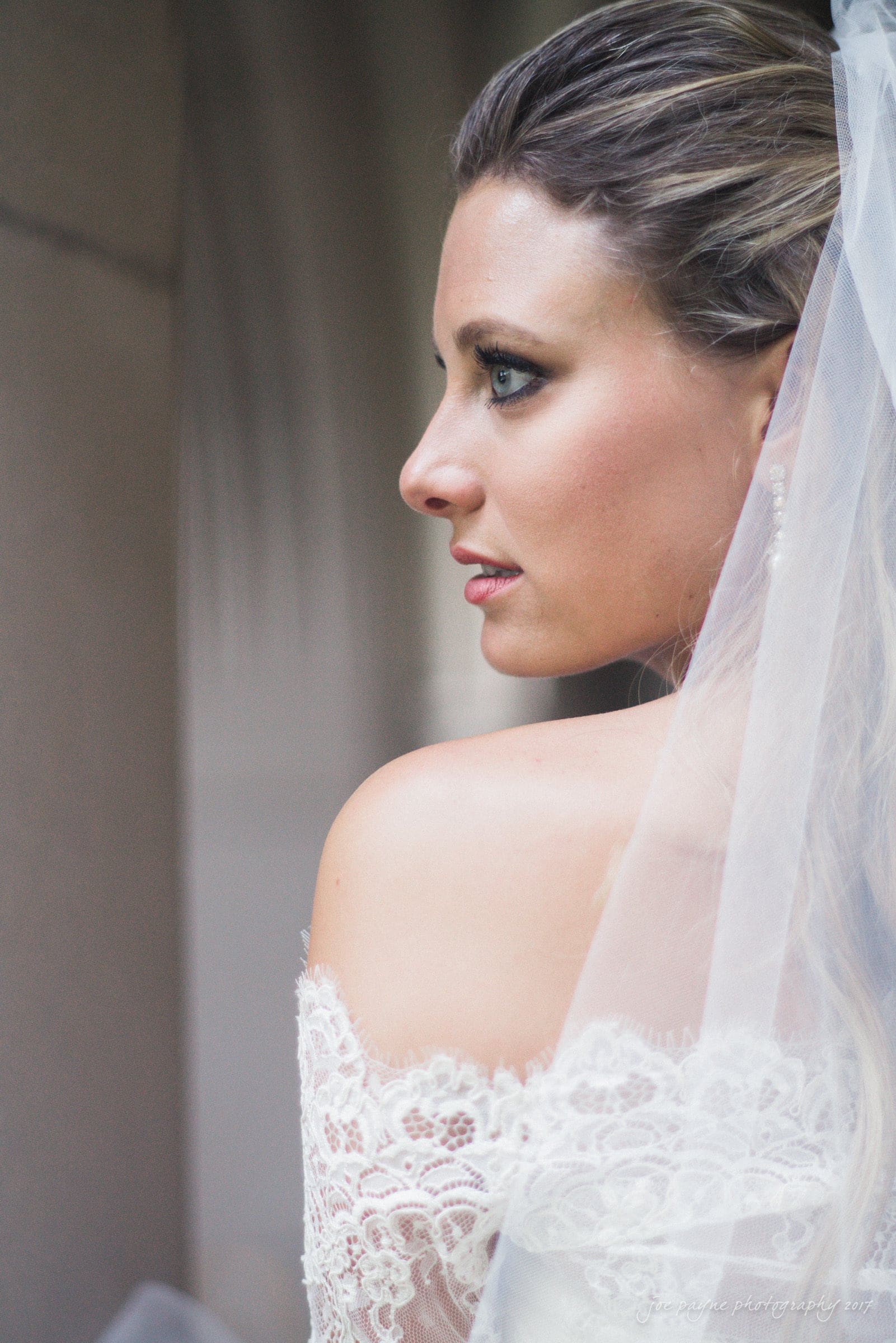 duke chapel wedding photography - sara's bridal session A bride in profile view, dressed in a lace bridal gown. Her hair is styled elegantly, and she wears a veil that drapes over her shoulders, highlighting her serene expression. The soft background gives a timeless appearance to the portrait.