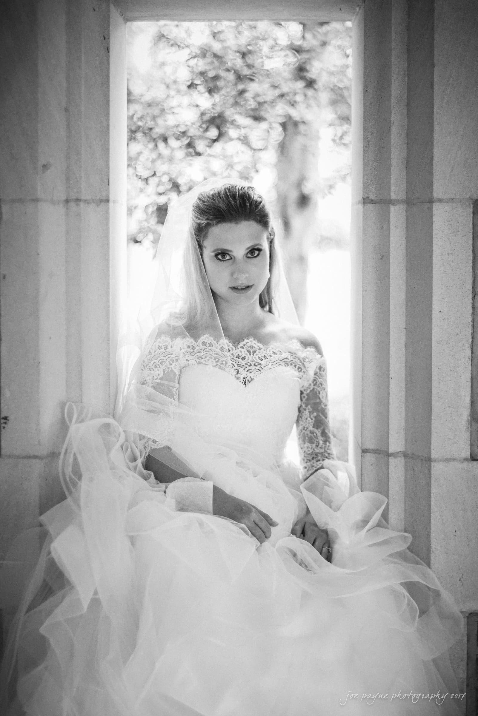 duke chapel wedding photography - sara's bridal session A bride in a lace wedding dress sits gracefully in a stone window frame. Her long veil and soft curls complement the elegant gown, while natural light creates a serene ambiance.
