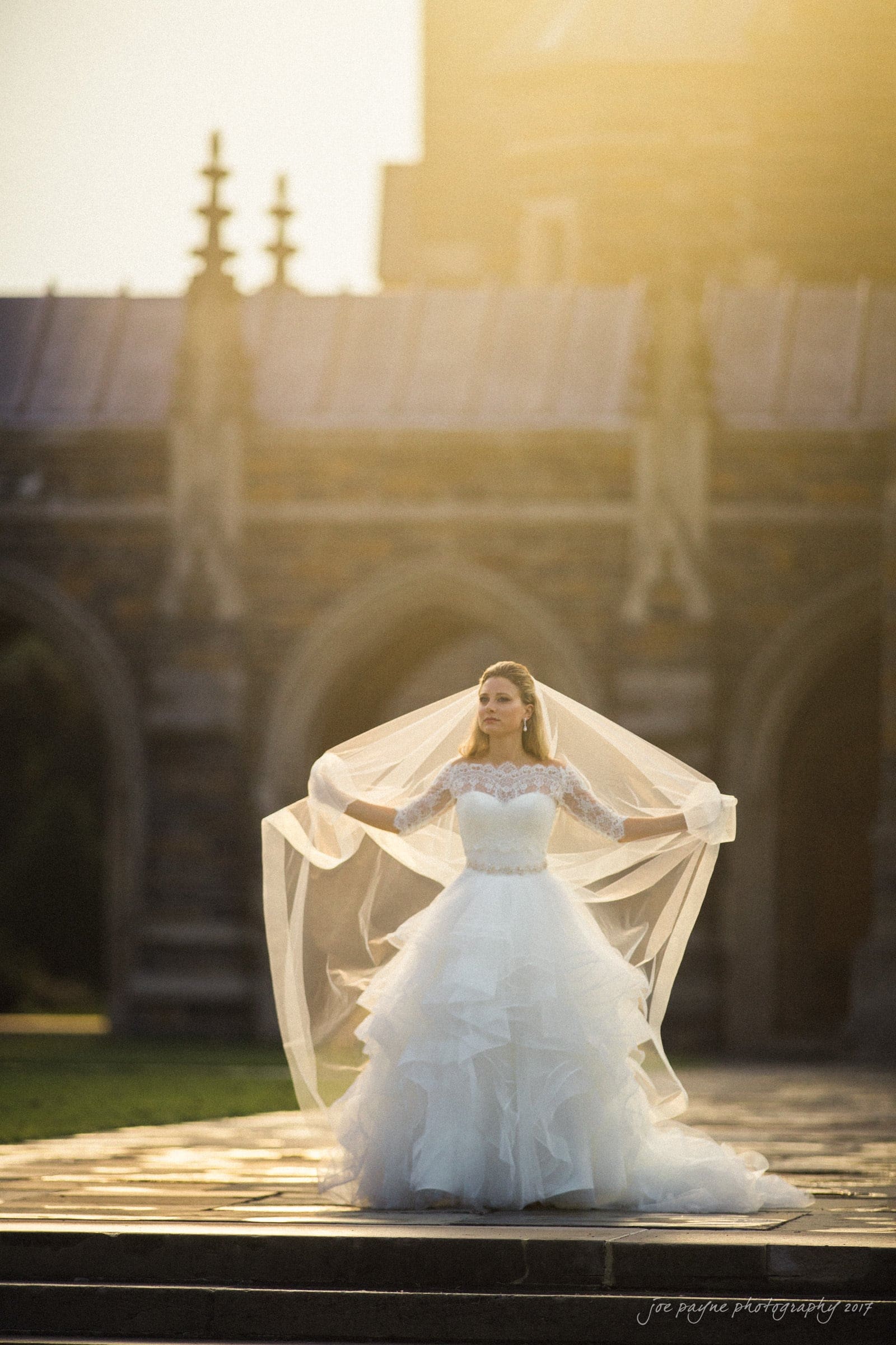 duke chapel wedding photography - sara's bridal session A bride in a flowing white wedding gown stands outdoors in front of a gothic-style building. She is holding a long, sheer veil with both hands, draping it around her. The scene is bathed in warm, golden sunlight.
