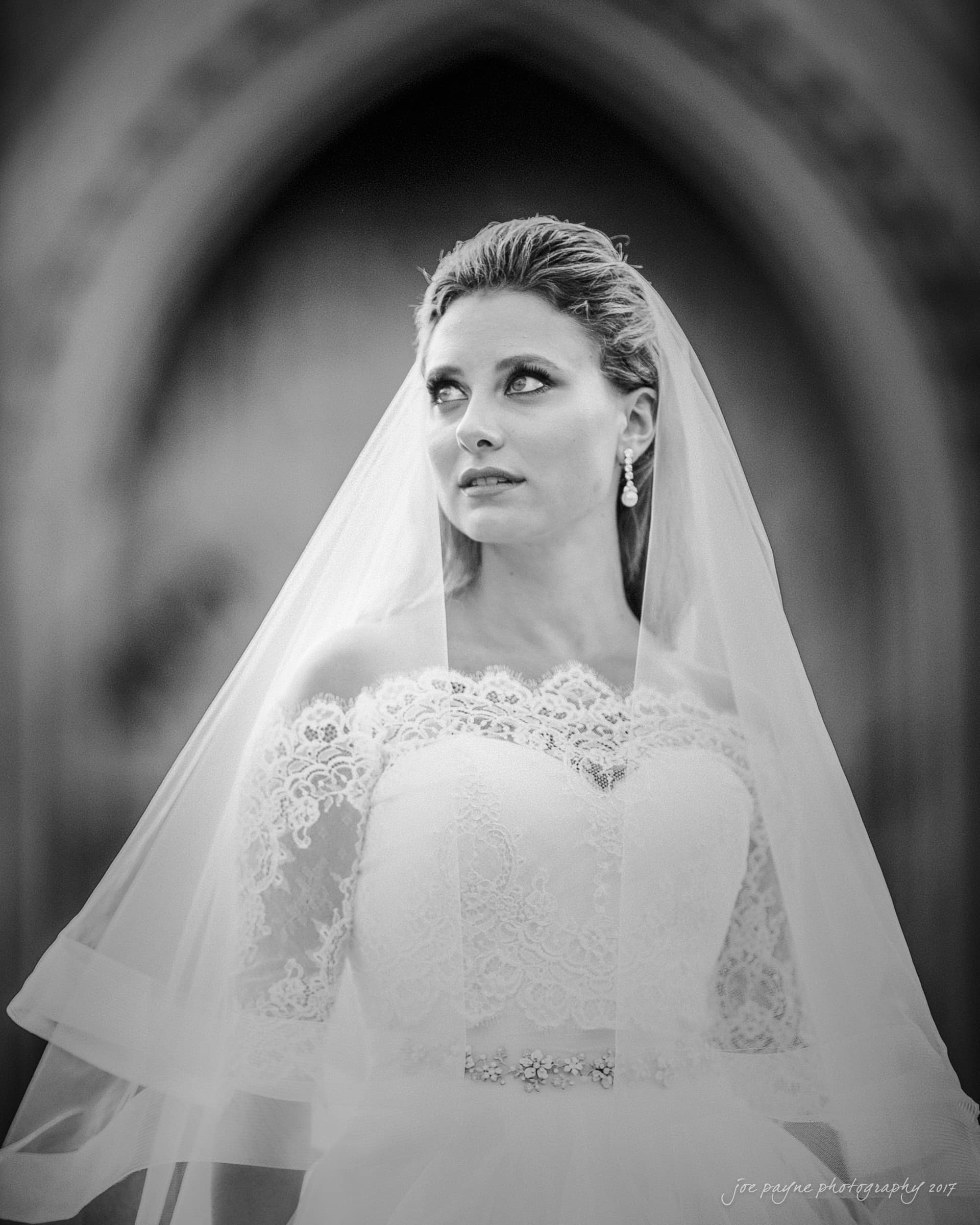 duke chapel wedding photography - sara's bridal session A bride in a lace wedding gown and veil gazes upwards, set against a blurred architectural background. The black and white image emphasizes her serene expression and elegant attire.
