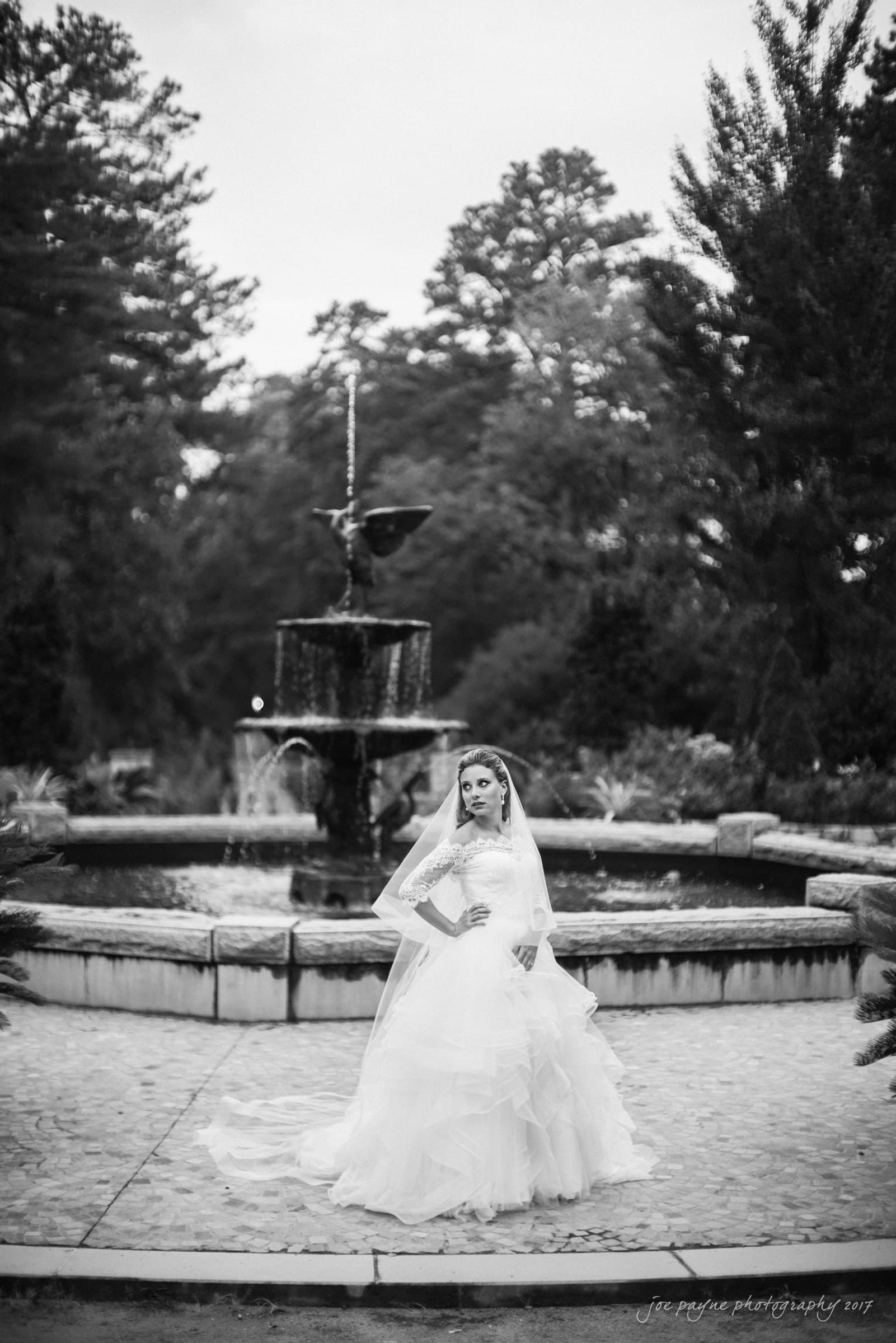 duke chapel wedding photography - sara's bridal session A bride in a flowing gown stands confidently in front of a tiered stone fountain. The outdoor setting is lush with tall trees and greenery, creating a serene and elegant atmosphere. The photo is in black and white.