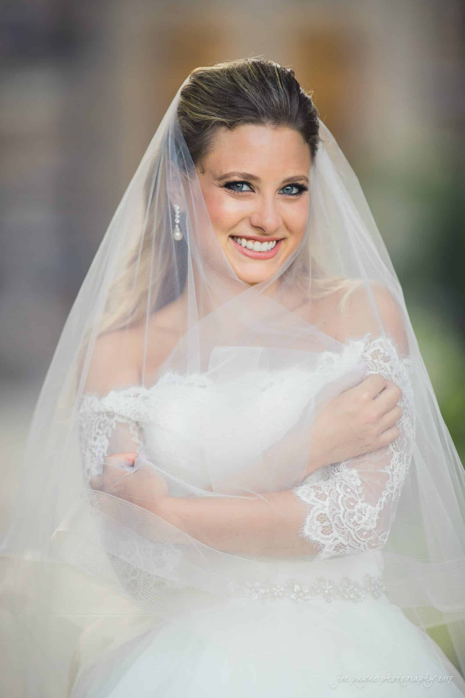 duke chapel wedding photography - sara's bridal session A bride smiles warmly, wearing a white wedding gown with lace details and a sheer veil draped over her shoulders. Her hair is styled elegantly, and she is standing outdoors, embracing the joyful moment.