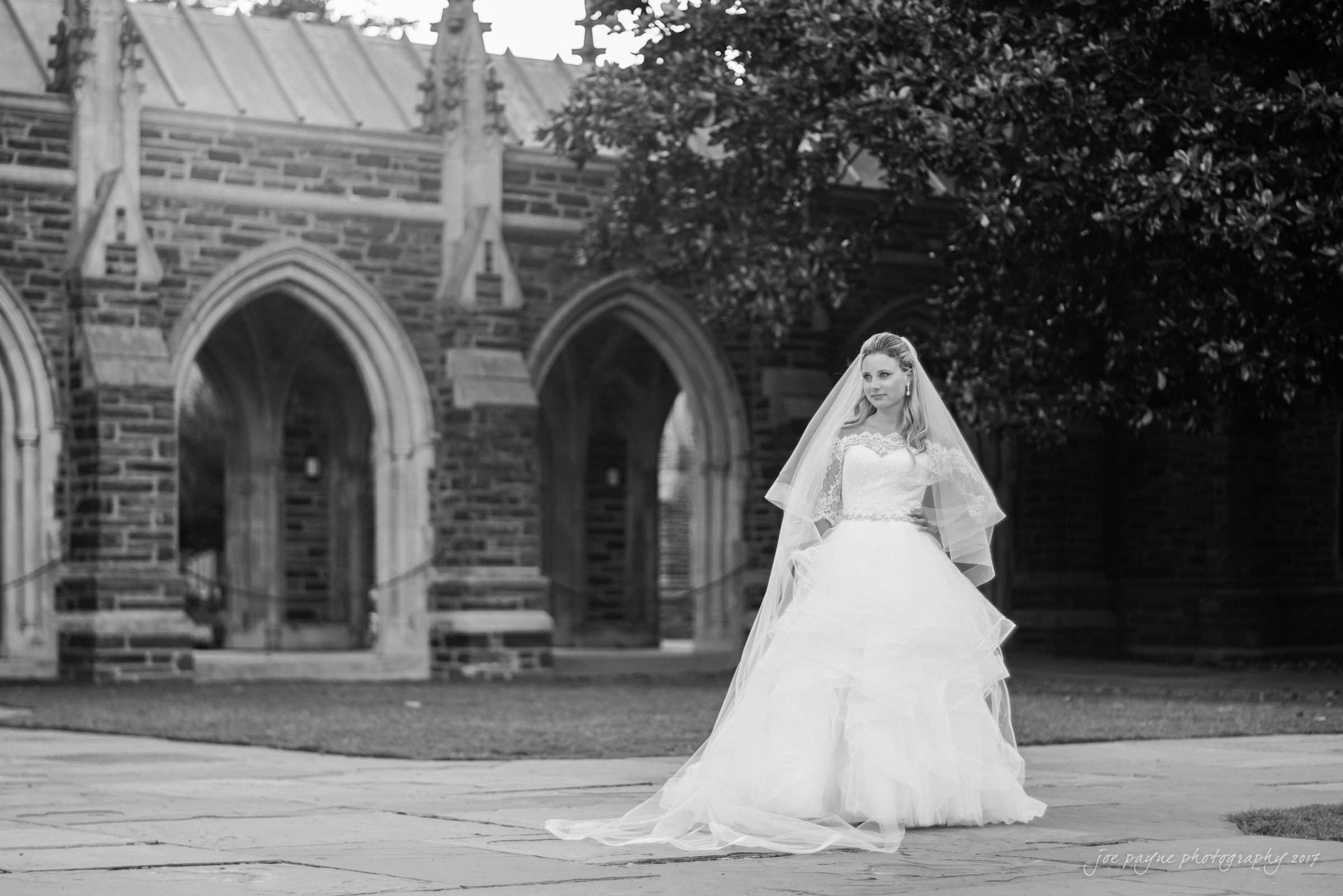 duke chapel wedding photography - sara's bridal session Black and white photo of a bride in a flowing wedding gown and veil, standing on a stone path. Arched stone building and trees are in the background, creating a serene and elegant atmosphere.