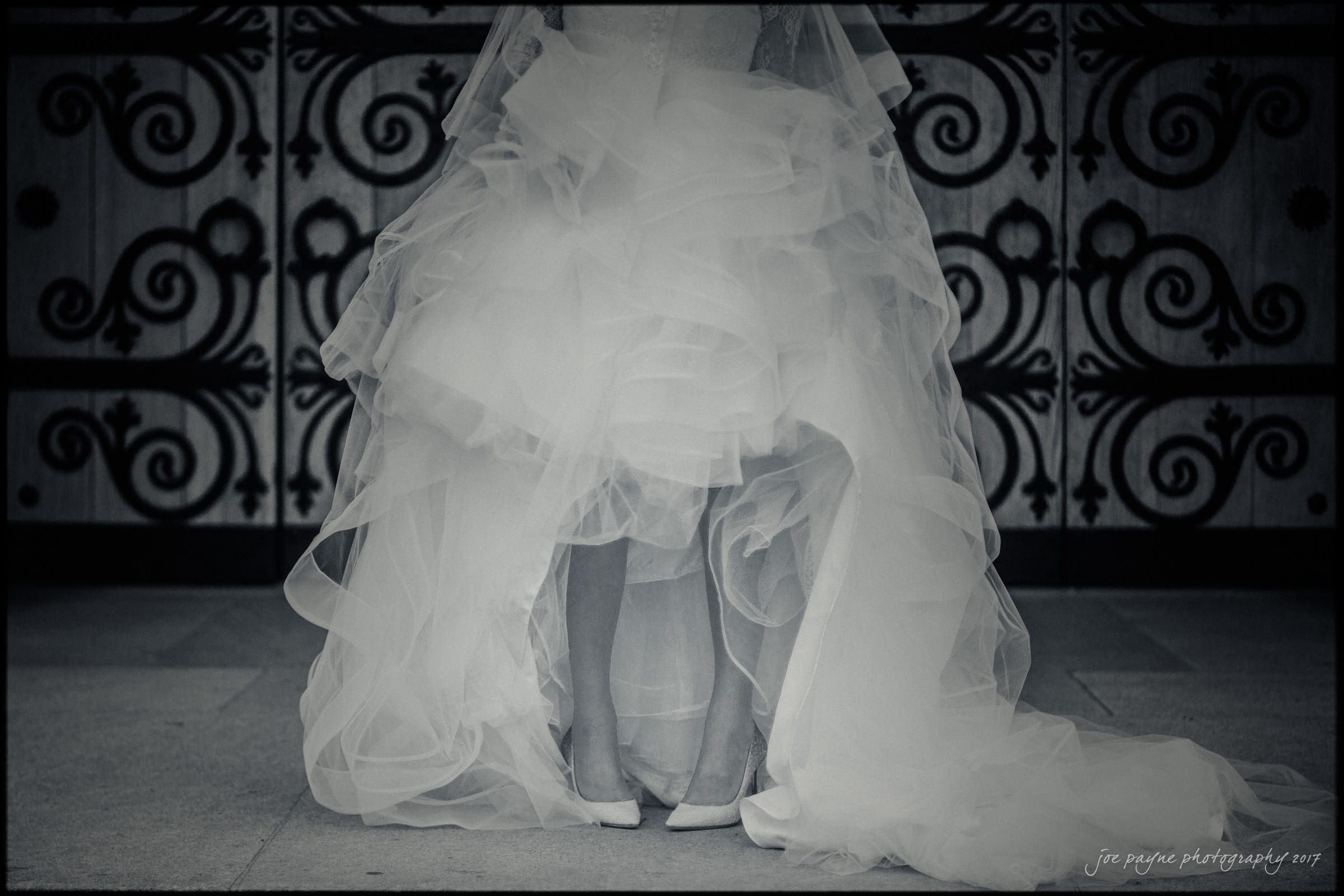 duke chapel wedding photography - sara's bridal session A black and white photo of a bride in a voluminous, ruffled wedding gown, revealing her feet in white shoes. The ornate, wrought-iron backdrop adds elegance to the composition.