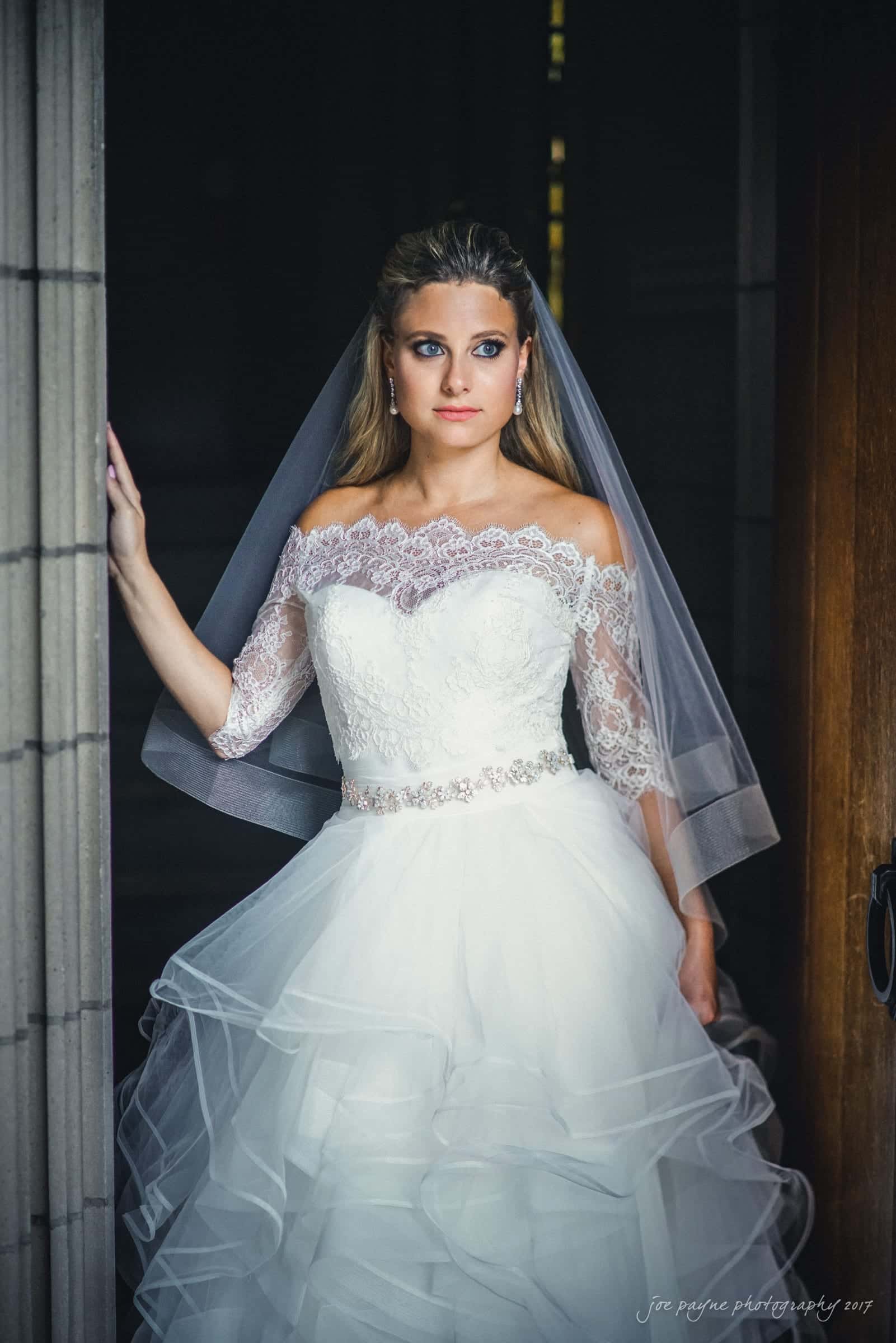 duke chapel wedding photography - sara's bridal session A bride stands in a doorway, wearing a white wedding dress with lace detail and an off-the-shoulder design. Her veil is draped over her shoulders, and she looks intently ahead. The background is dark, accentuating her presence.