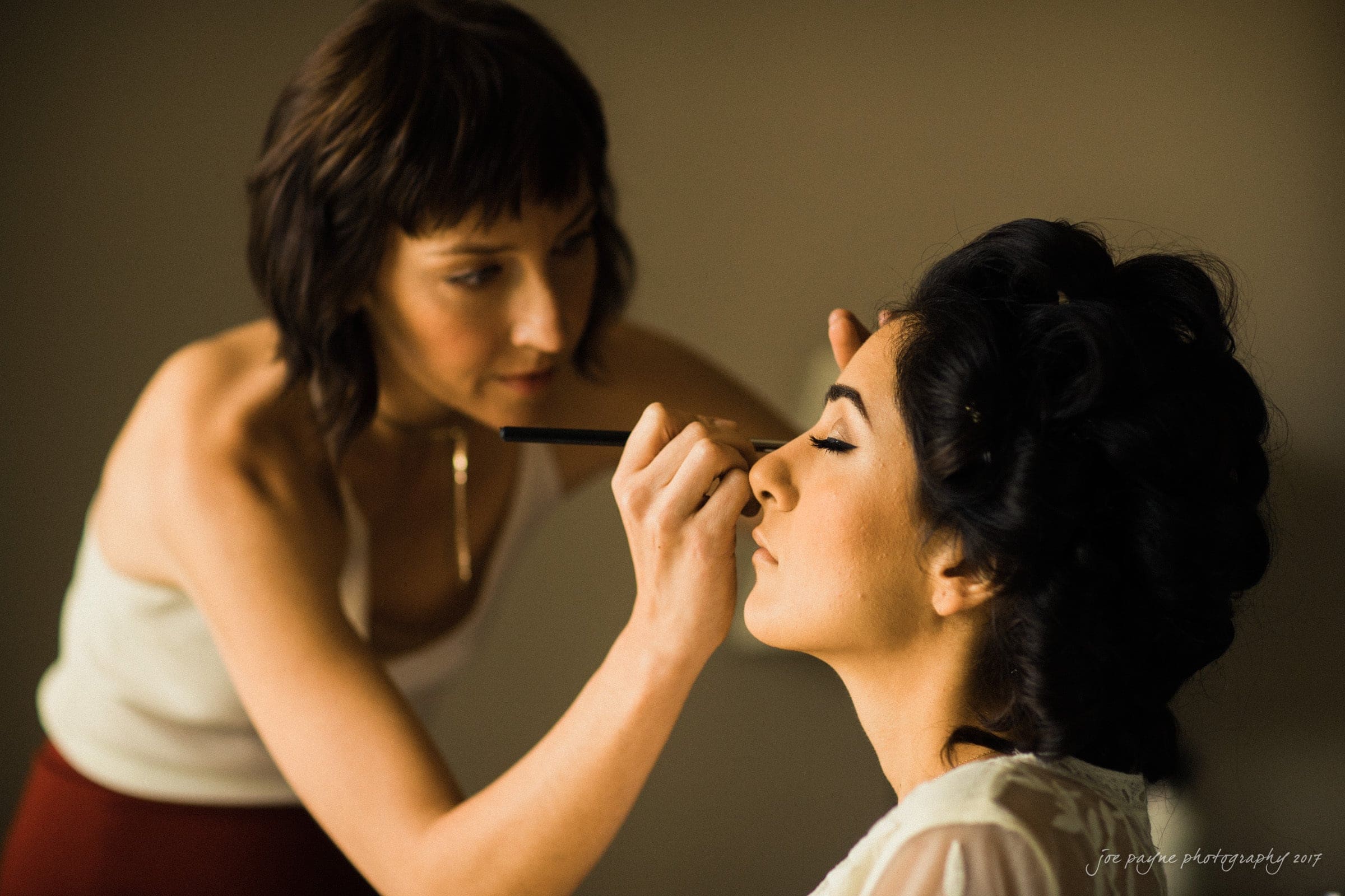 A woman with short dark hair carefully applies makeup to another womans face. The woman receiving makeup has her eyes closed and dark, curled hair. The setting appears calm and focused.