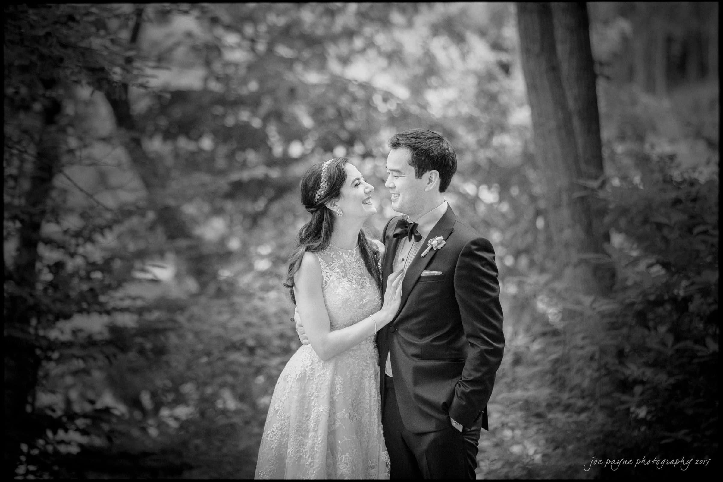A bride and groom stand close together outdoors, smiling and gazing at each other lovingly. Captured by Umstead wedding photography, the bride wears a sleeveless lace gown while the groom is in a suit, surrounded by lush trees and foliage.