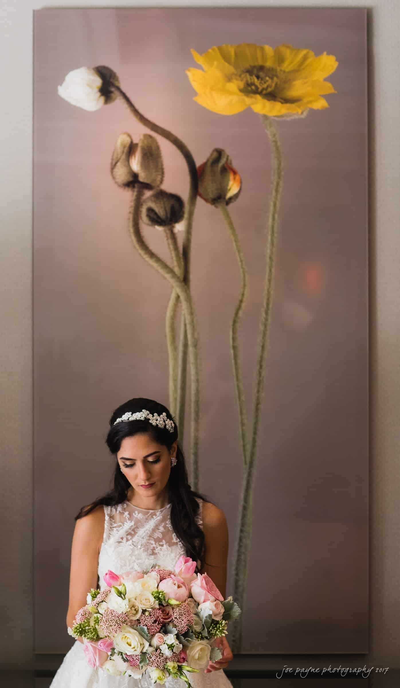A bride in a white lace dress holds a bouquet of pink and white flowers. She stands in front of a large artwork depicting tall yellow and white flowers.