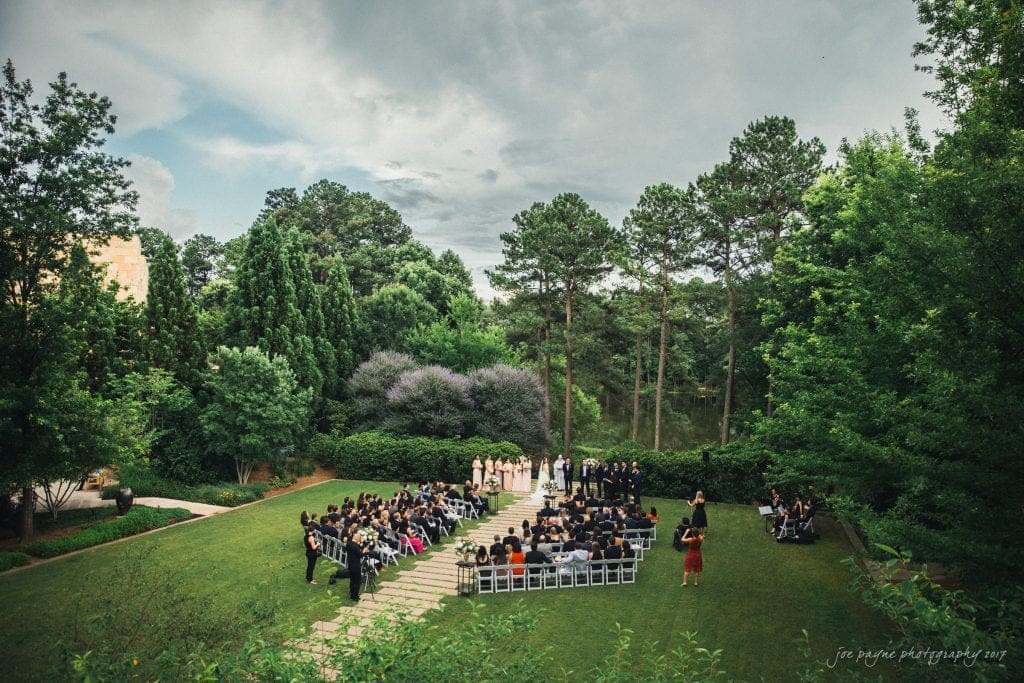 A wedding ceremony is taking place in a lush outdoor setting surrounded by trees. Guests are seated in rows of chairs facing the couple at the altar. The sky is partly cloudy, creating a serene and intimate atmosphere.