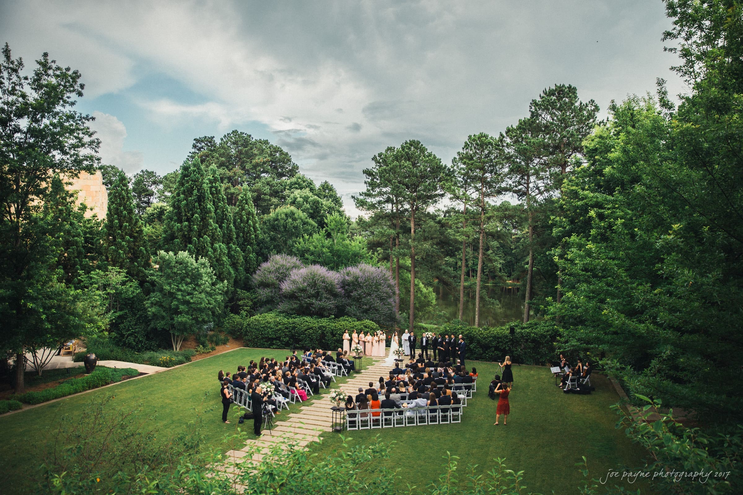 A wedding ceremony is taking place in a lush outdoor setting surrounded by trees. Guests are seated in rows of chairs facing the couple at the altar. The sky is partly cloudy, creating a serene and intimate atmosphere.