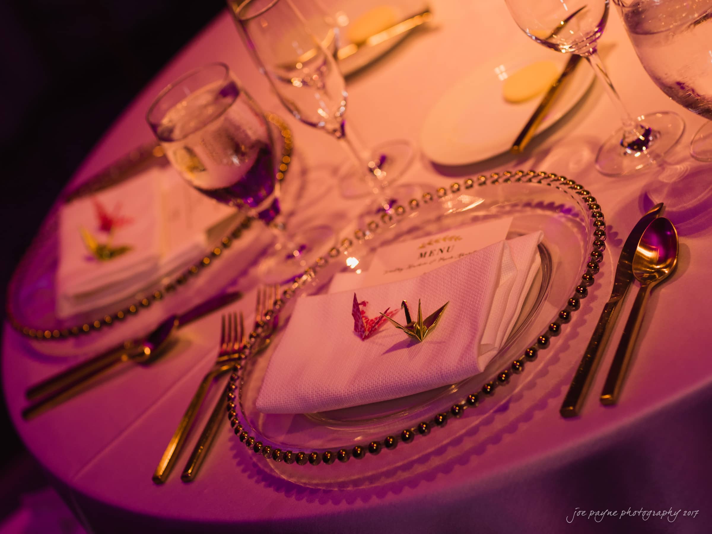 A table is elegantly set with two clear glass plates on white napkins, featuring pink floral decorations. Surrounding the plates, theres gold cutlery and wine glasses, all bathed in soft purple lighting.