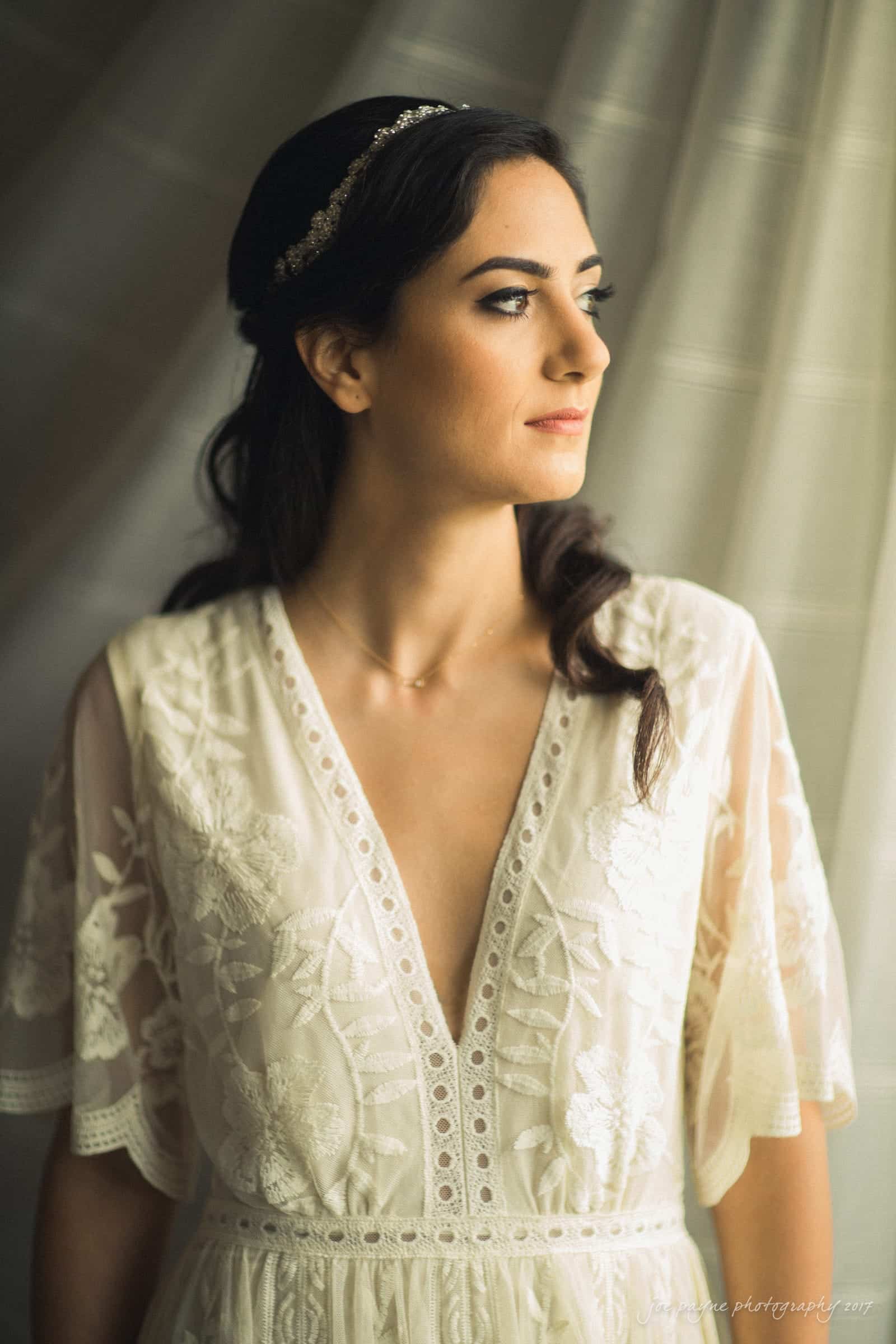 A woman in a white lace dress and headband stands by a window, gazing outside. The soft lighting accentuates her dresss intricate floral patterns, creating an elegant and serene atmosphere.