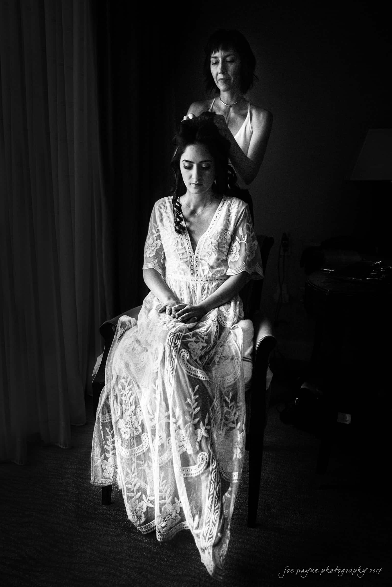 A black and white image shows a woman seated, wearing an intricately designed lace dress, while another woman styles her hair. The scene is illuminated softly, creating a serene and intimate atmosphere.