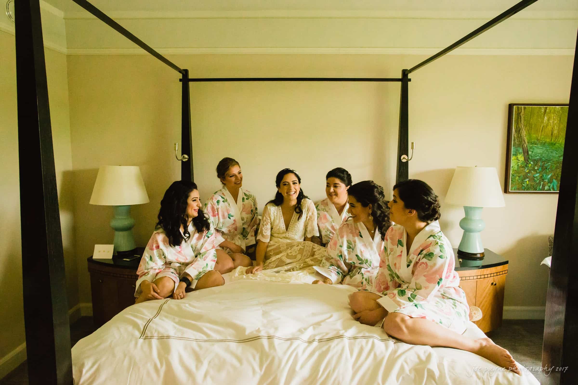 A group of six women, wearing floral robes, sits together on a large four-poster bed in a warmly lit room. They are engaged in conversation, smiling, and sharing a joyful moment. A painting and lamps are visible in the background.