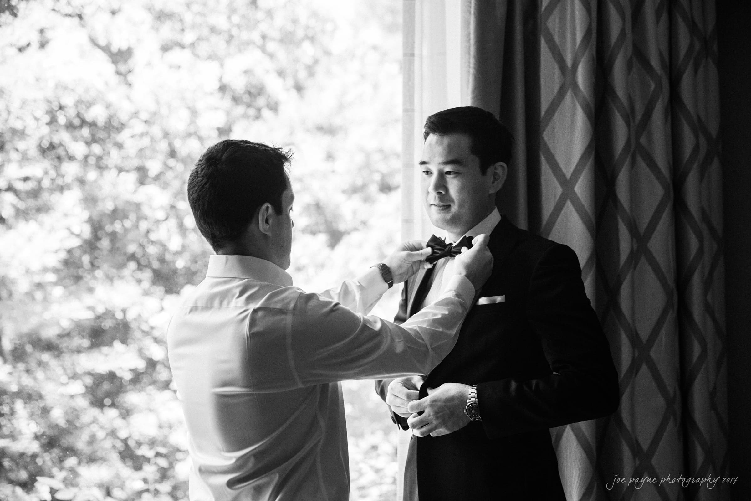 A man helps another man adjust his bow tie in front of a window. The scene is in black and white, with the two men dressed formally in a suit and a shirt. Curtains and blurred outdoor foliage are in the background.