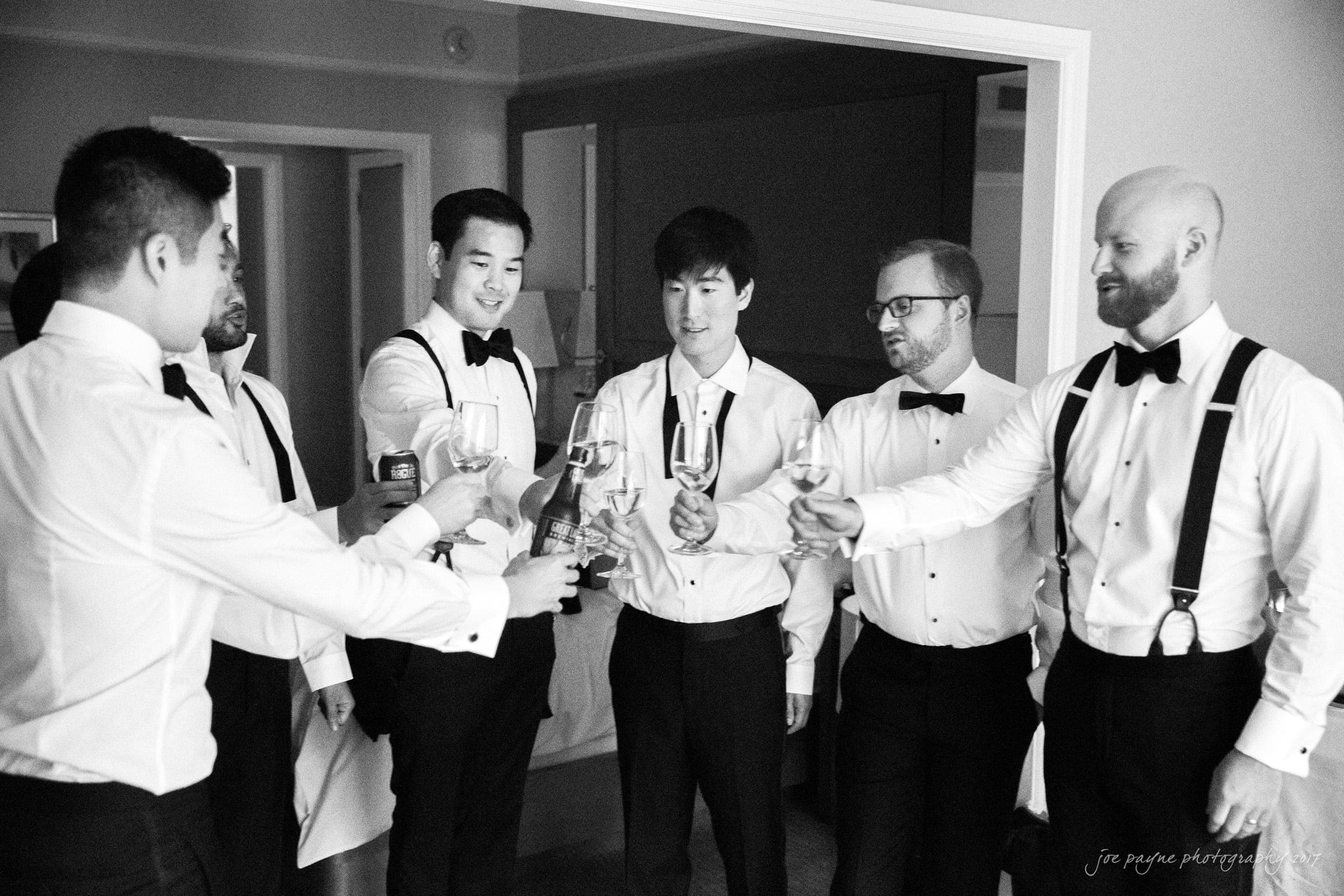 A group of six men in formal attire, including shirts, bow ties, and suspenders, raise their glasses in a celebratory toast in a room. The mood is joyful and relaxed. The photo is in black and white.