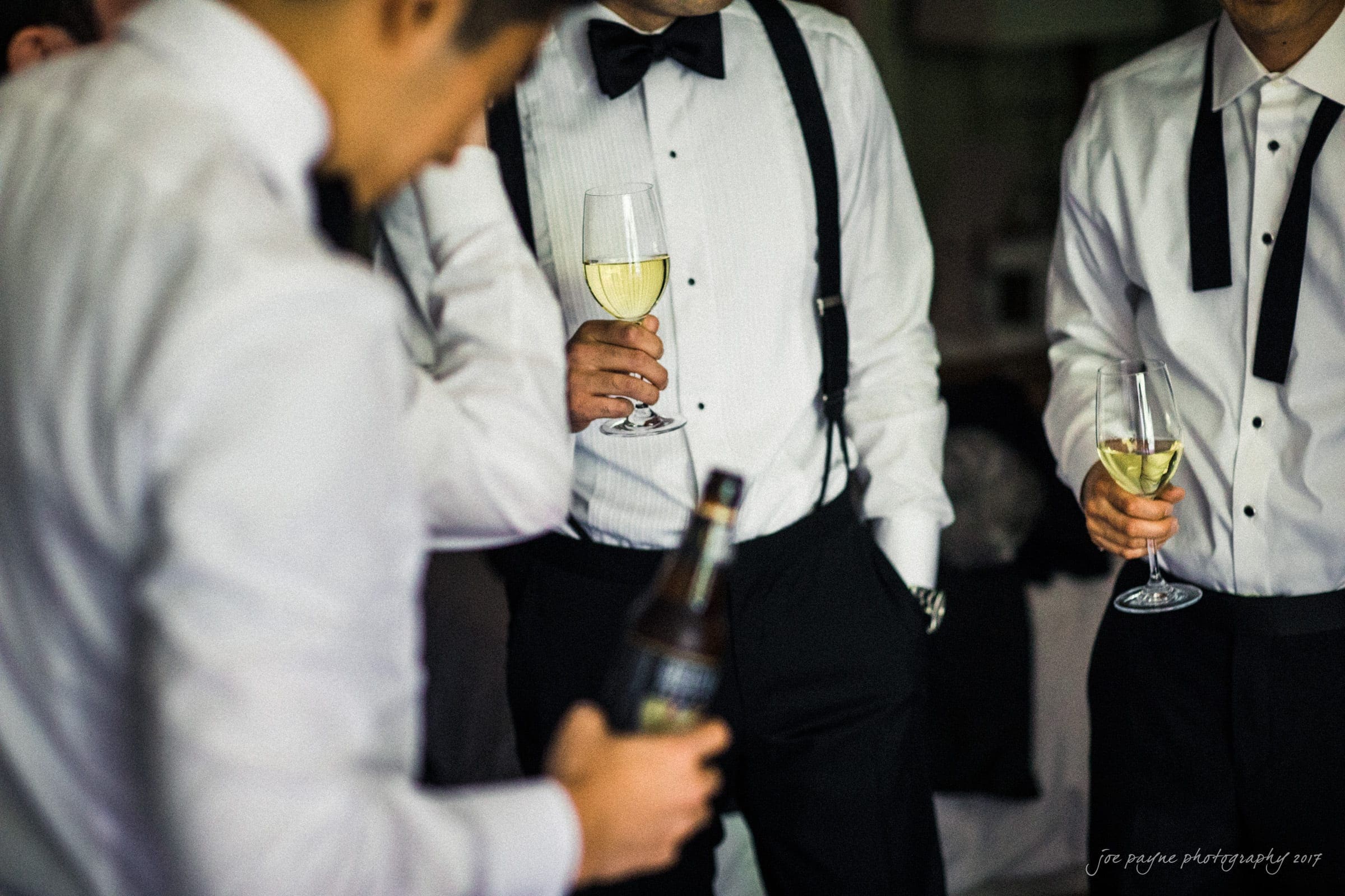 A group of men in formal attire, wearing white shirts, black bow ties, and suspenders, holding glasses of champagne. One is holding a bottle. The setting appears to be a social gathering or celebration.