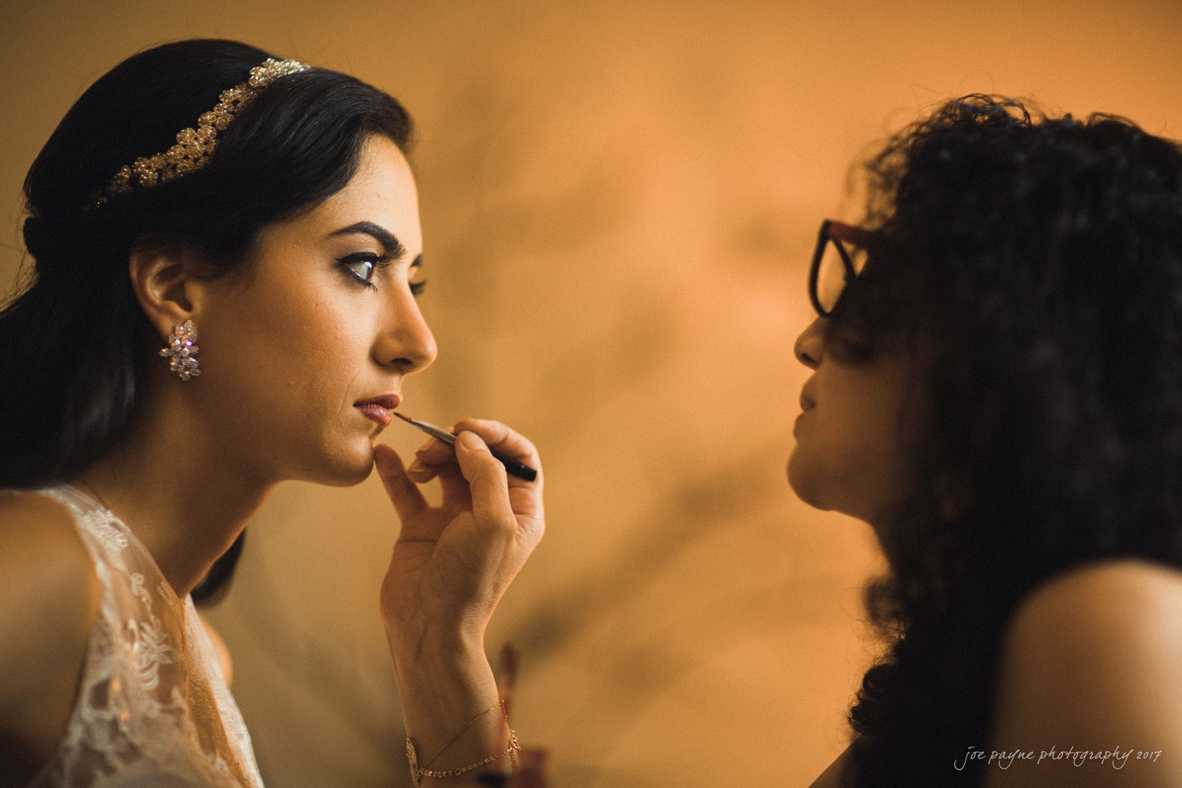 A bride with long dark hair and a jeweled headband is having her makeup applied by a woman with curly hair and glasses. The bride is dressed in white, and the scene is warmly lit, creating an intimate atmosphere.