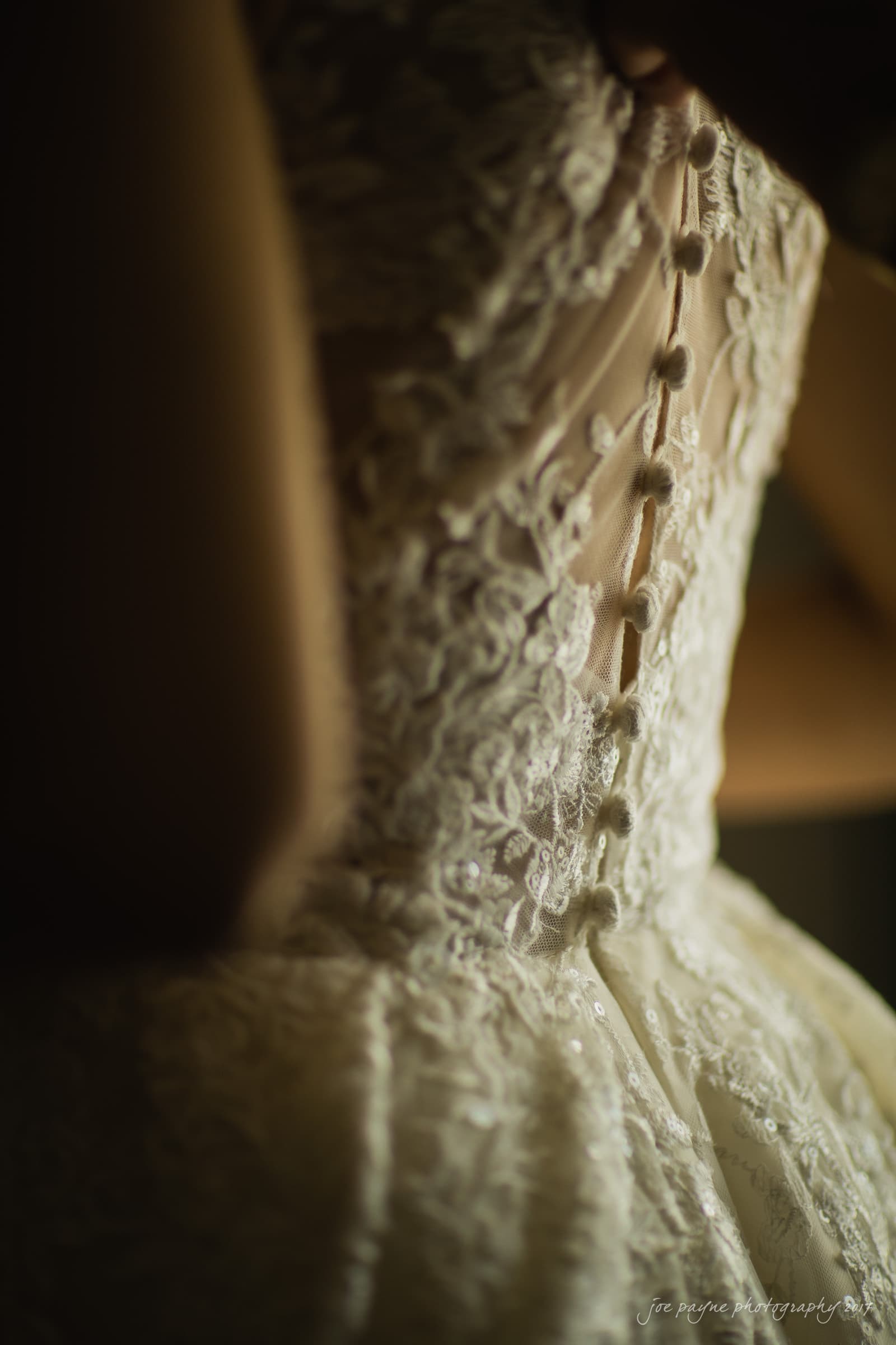 Close-up of a person wearing a lace wedding dress, focusing on the intricate button details on the bodice. The image captures the delicate texture and artistry of the fabric under soft lighting.