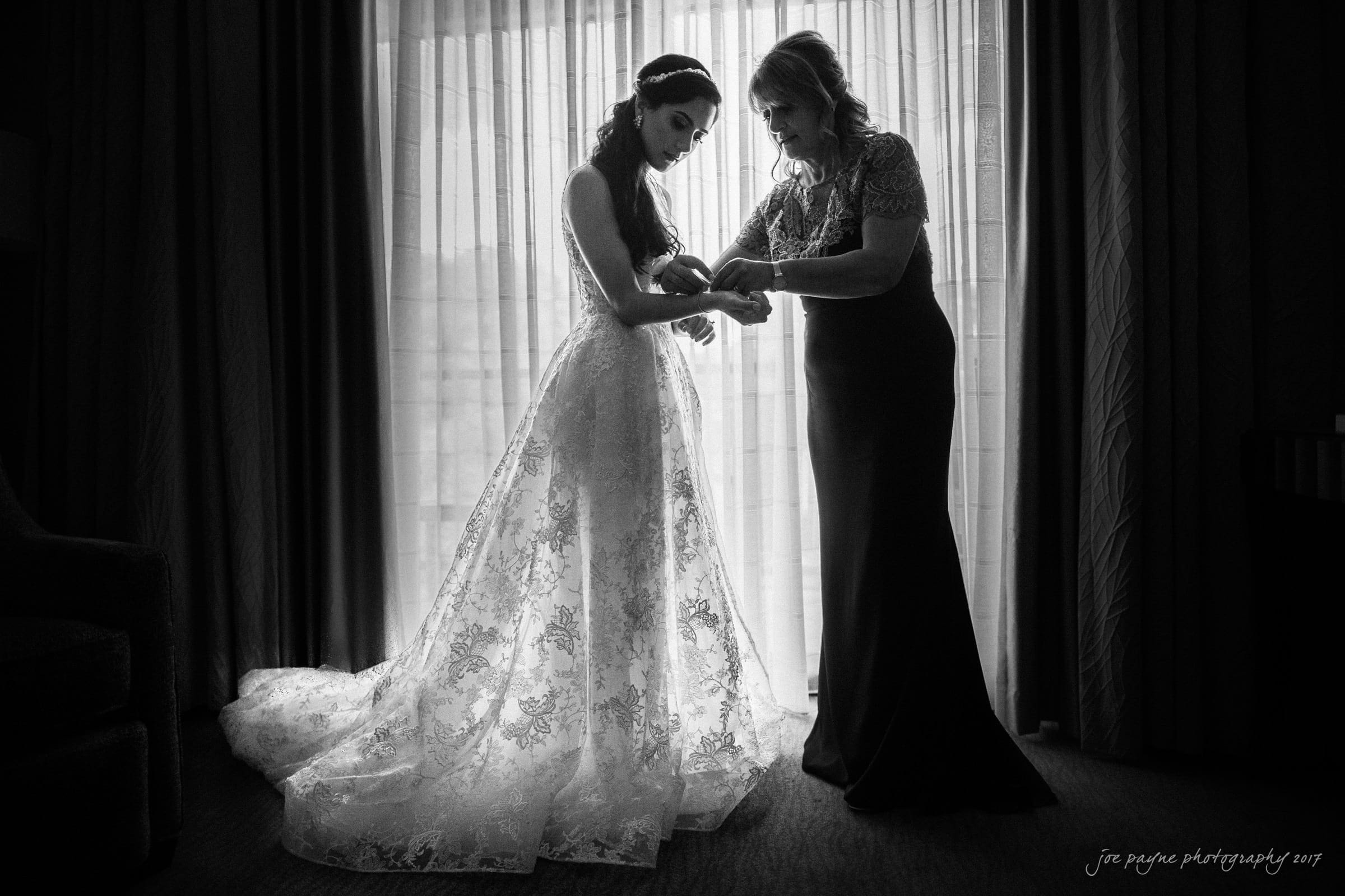 A bride in a lace wedding gown stands in front of a window as a woman in an elegant dress helps adjust her bracelet. The scene is black and white, with light filtering through sheer curtains, creating a soft, intimate atmosphere.