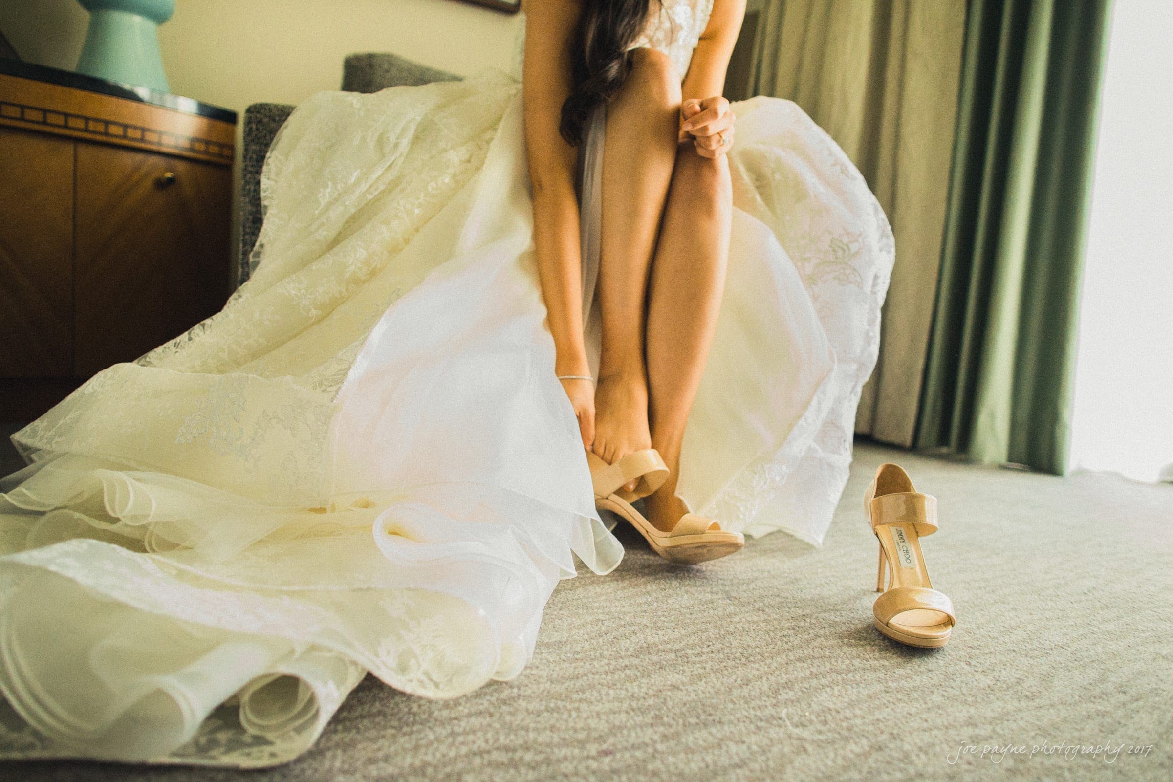 A bride sitting on a chair adjusts her beige high-heeled shoe. She wears a lace wedding dress with a full skirt that cascades to the carpeted floor. Another shoe lies on the floor beside her. The room is softly lit.