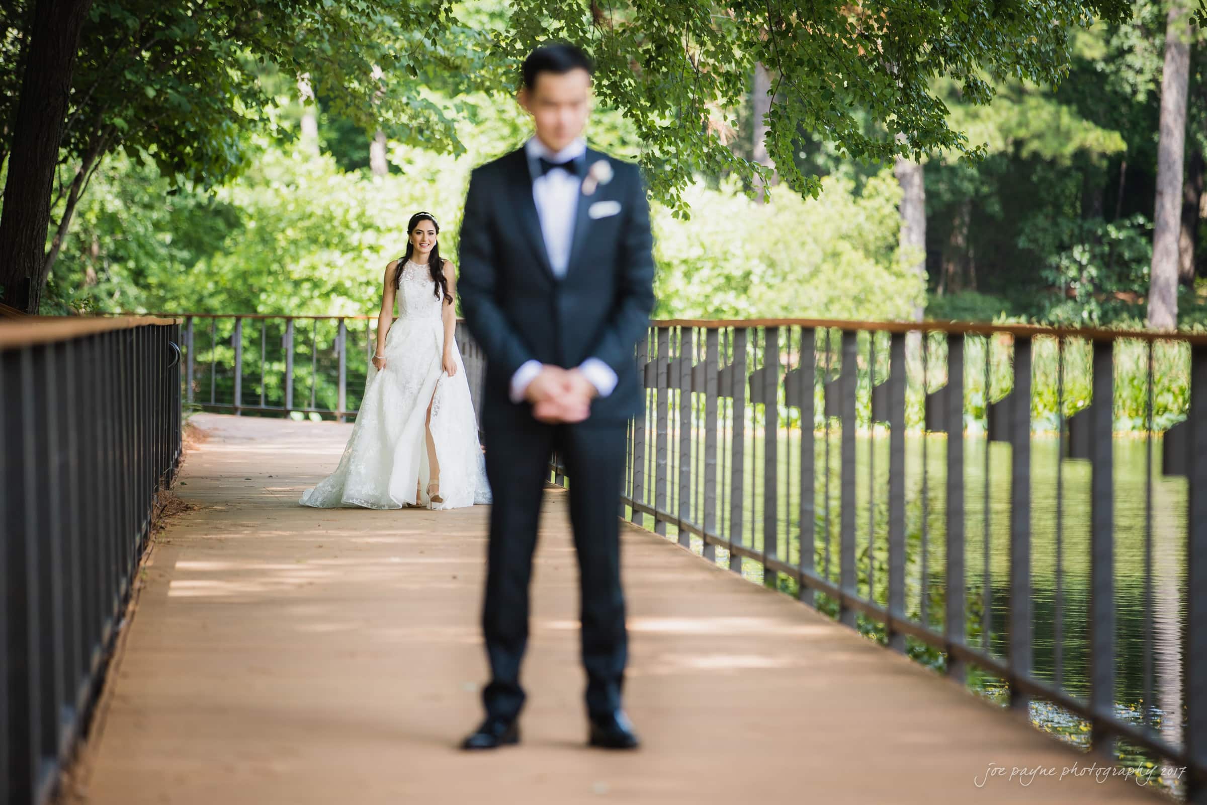A bride and groom are on a wooden walkway surrounded by trees. The groom, in a black suit, stands in the foreground with his back slightly to the camera, while the bride, in a white gown, approaches him from behind.