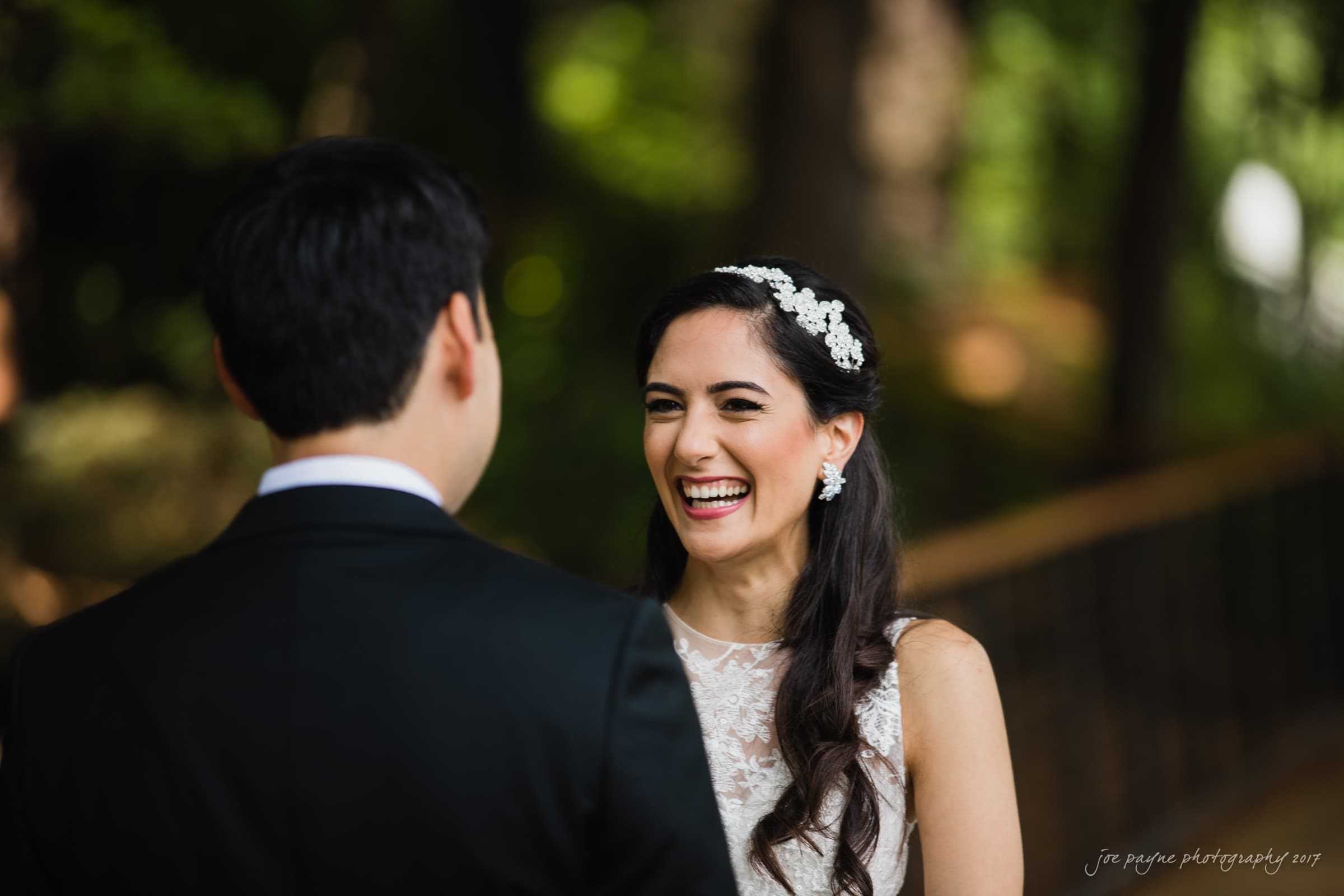 A woman in a white dress with floral accessories in her hair smiles at a man in a black suit. They are outdoors with greenery and a blurred background.