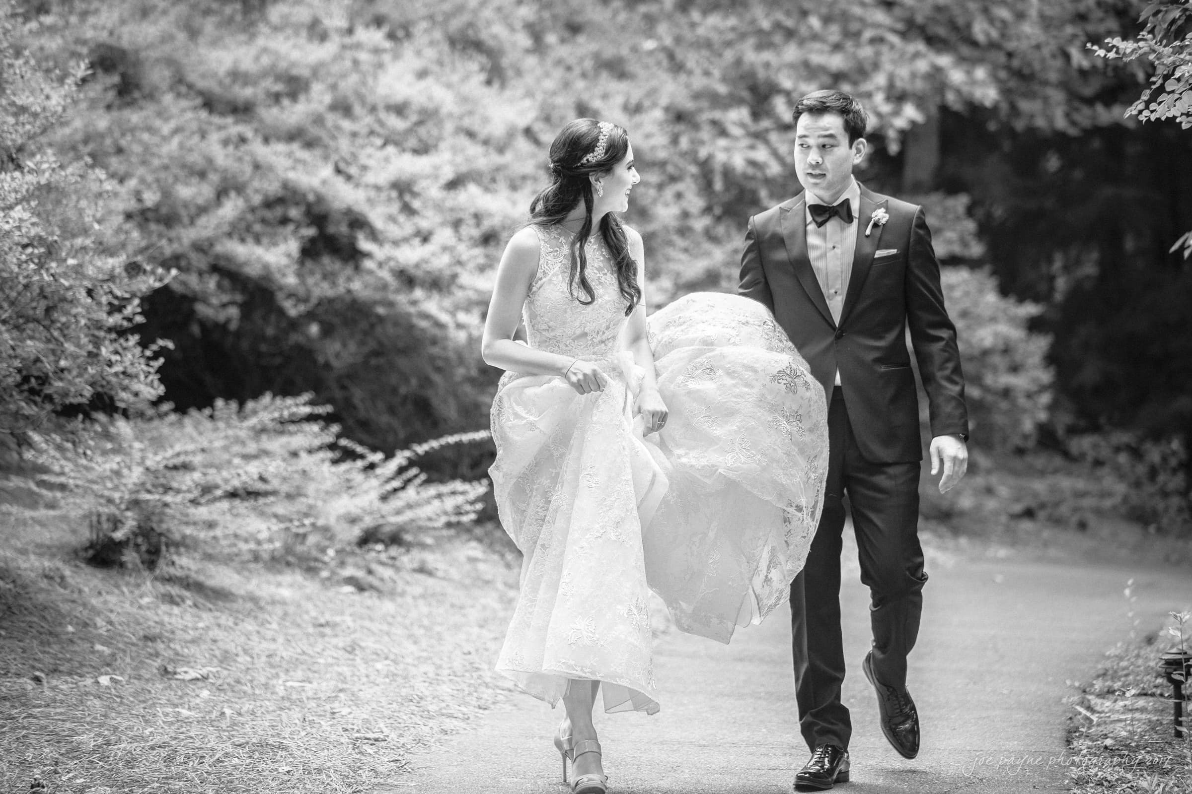 A black and white photo of a bride in a lace gown holding her dress, walking with a groom in a suit on a tree-lined path. Both are smiling, suggesting a joyful moment.