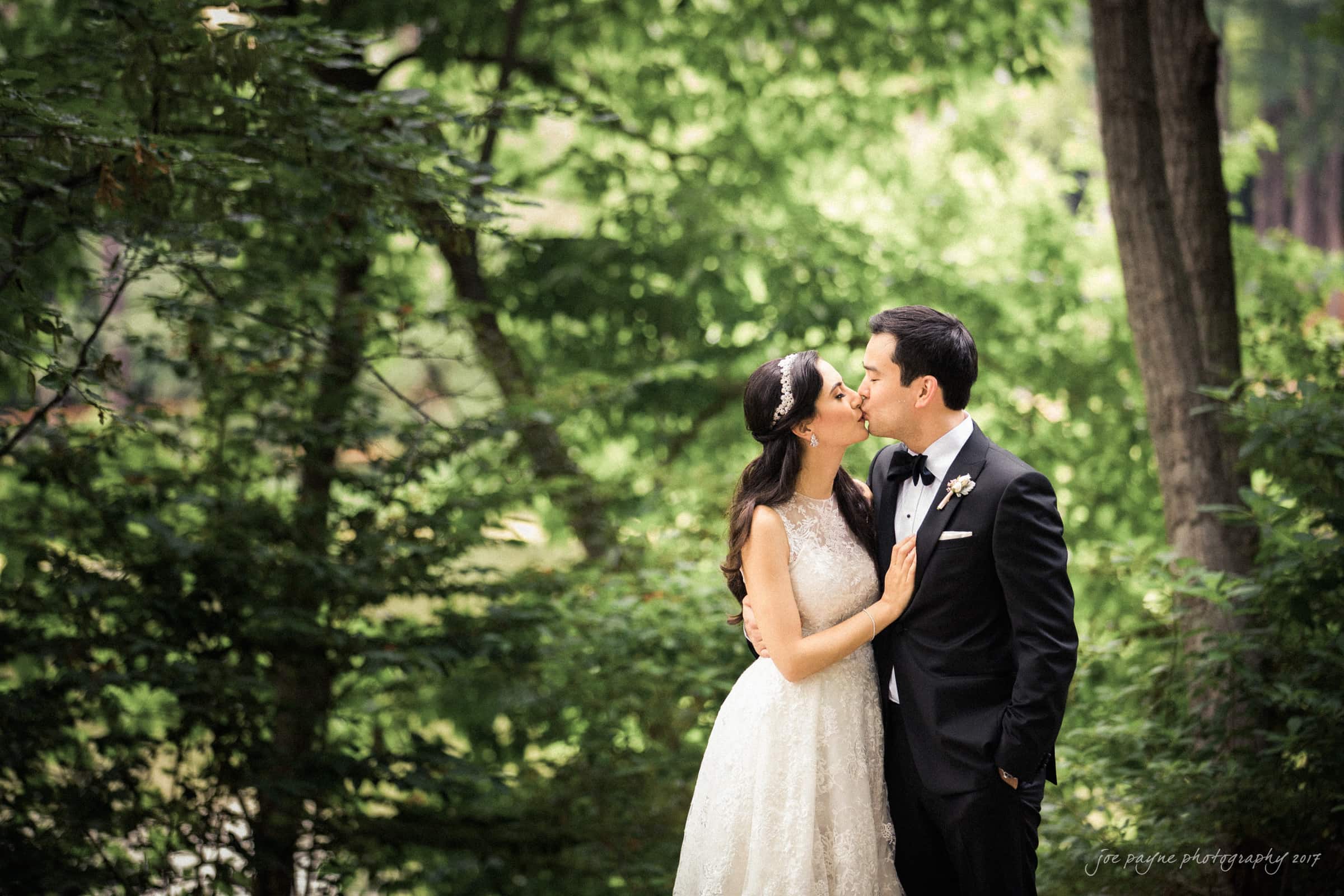 A bride and groom share a kiss in a lush, green forest setting. She wears a white lace gown and a decorative headpiece, while he is dressed in a black suit with a bow tie.