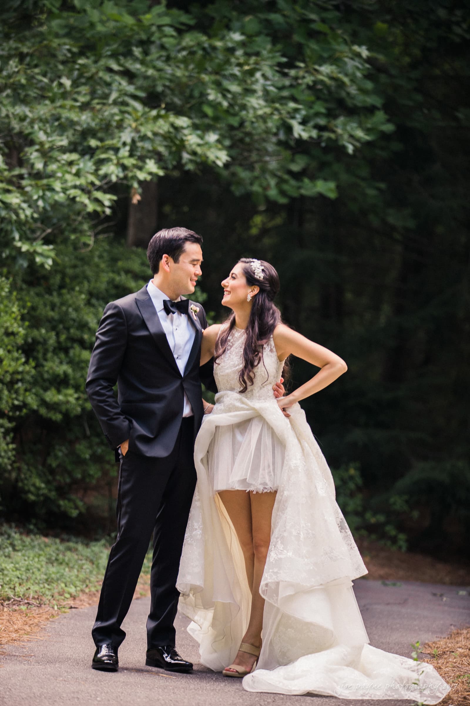 A bride and groom stand on a path surrounded by lush greenery. The groom wears a black tuxedo, and the bride wears a white high-low wedding dress with a long train. They are smiling and looking at each other.