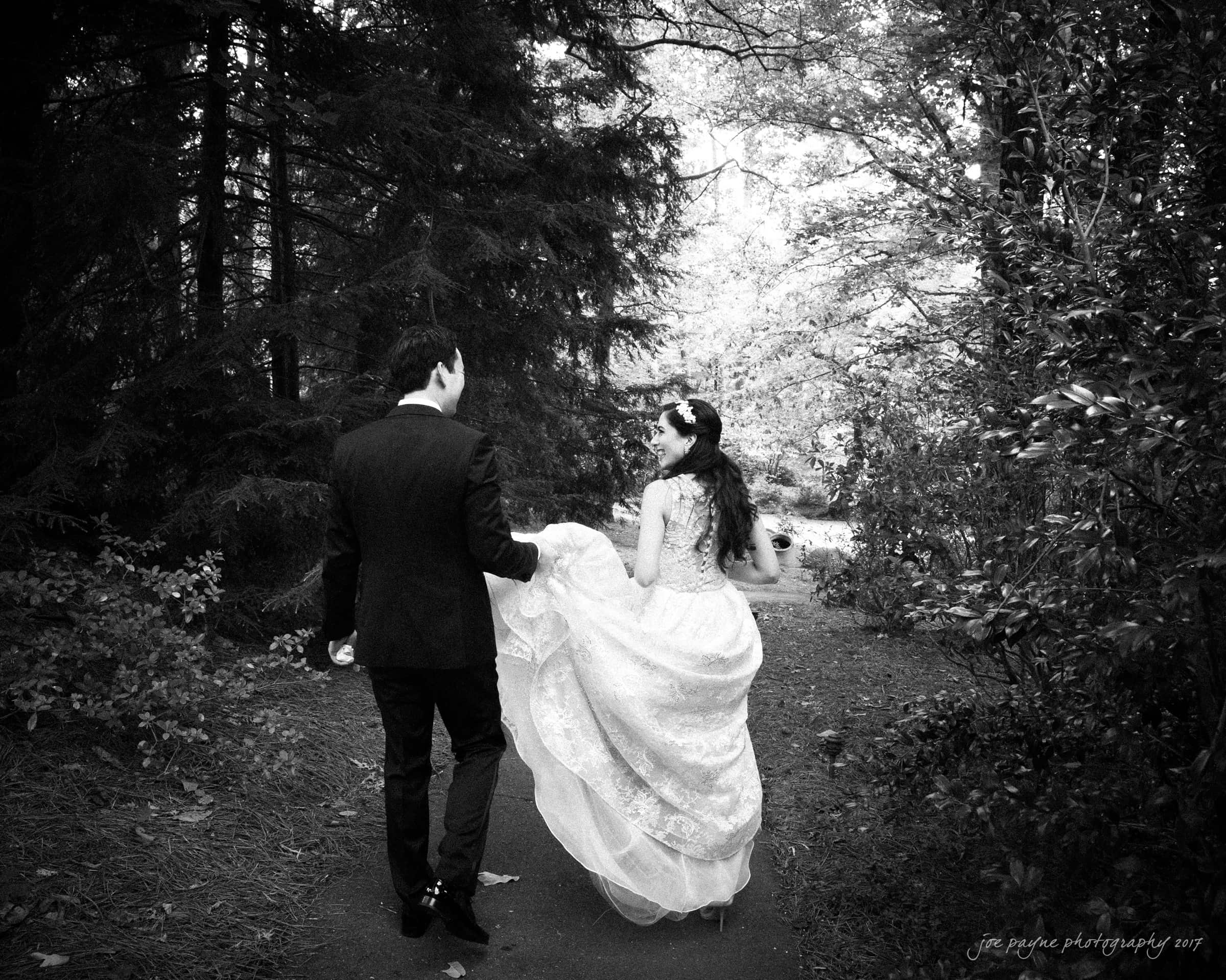 A black and white photo of a couple walking down a forest path. The woman is wearing a lace wedding dress, holding it up slightly, while the man is in a dark suit. They are walking away from the camera, surrounded by dense trees.