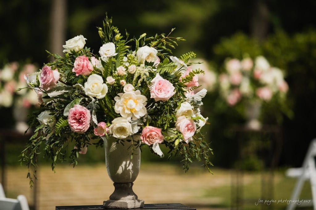 A vase filled with pink and white roses, greenery, and other flowers is displayed outdoors with a blurred background of similar floral arrangements.